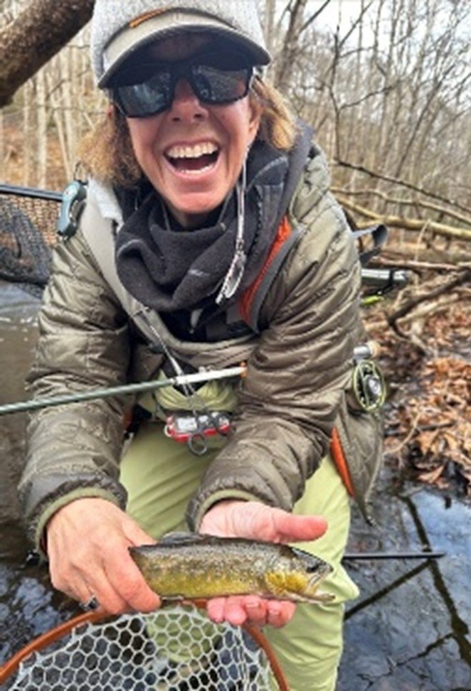Woman wearing sunglasses, a hat, and outdoor gear, smiling and holding a small fish with a net at her feet, in a wooded stream.