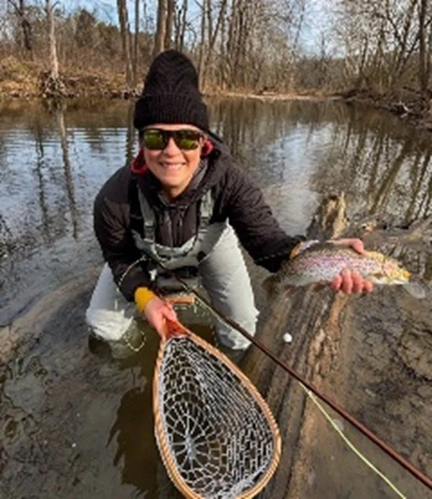 Person in waders, wearing sunglasses and a beanie, holding a large rainbow trout and a fishing net while kneeling in a shallow river.