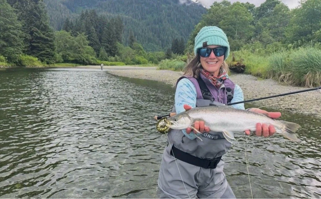 A woman in outdoor fishing gear holding a large fish she caught by a river in a forested area.