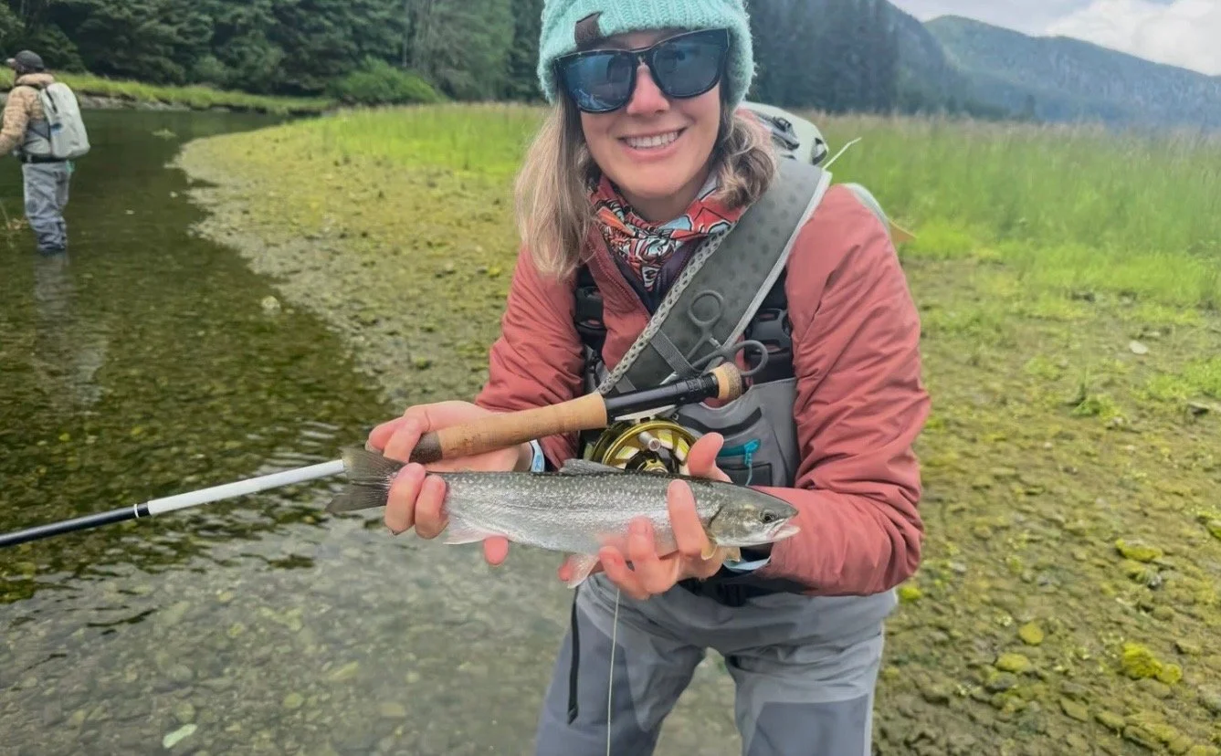 A woman smiling and holding a fish she caught while fishing in a river, wearing sunglasses, a beanie, and outdoor gear, with another person fishing in the background.