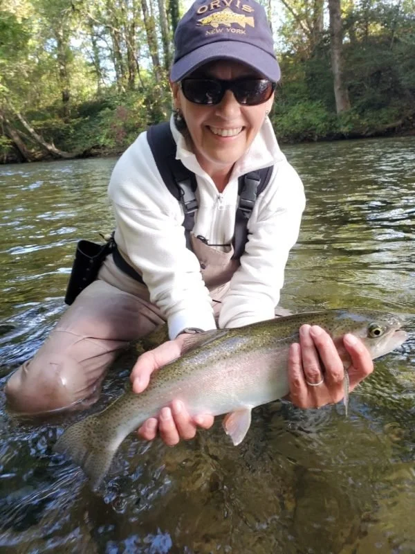 Woman wearing sunglasses, a cap, and outdoor gear, smiling and holding a large fish in a river surrounded by trees.
