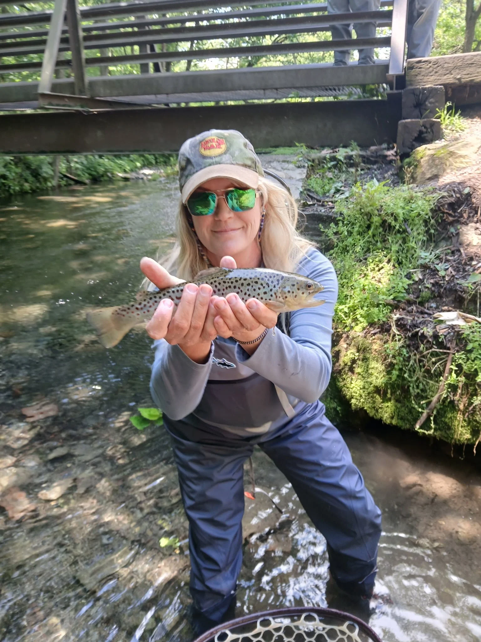 Woman holding a caught trout in a stream, wearing sunglasses and a cap, with a bridge and green foliage in the background.