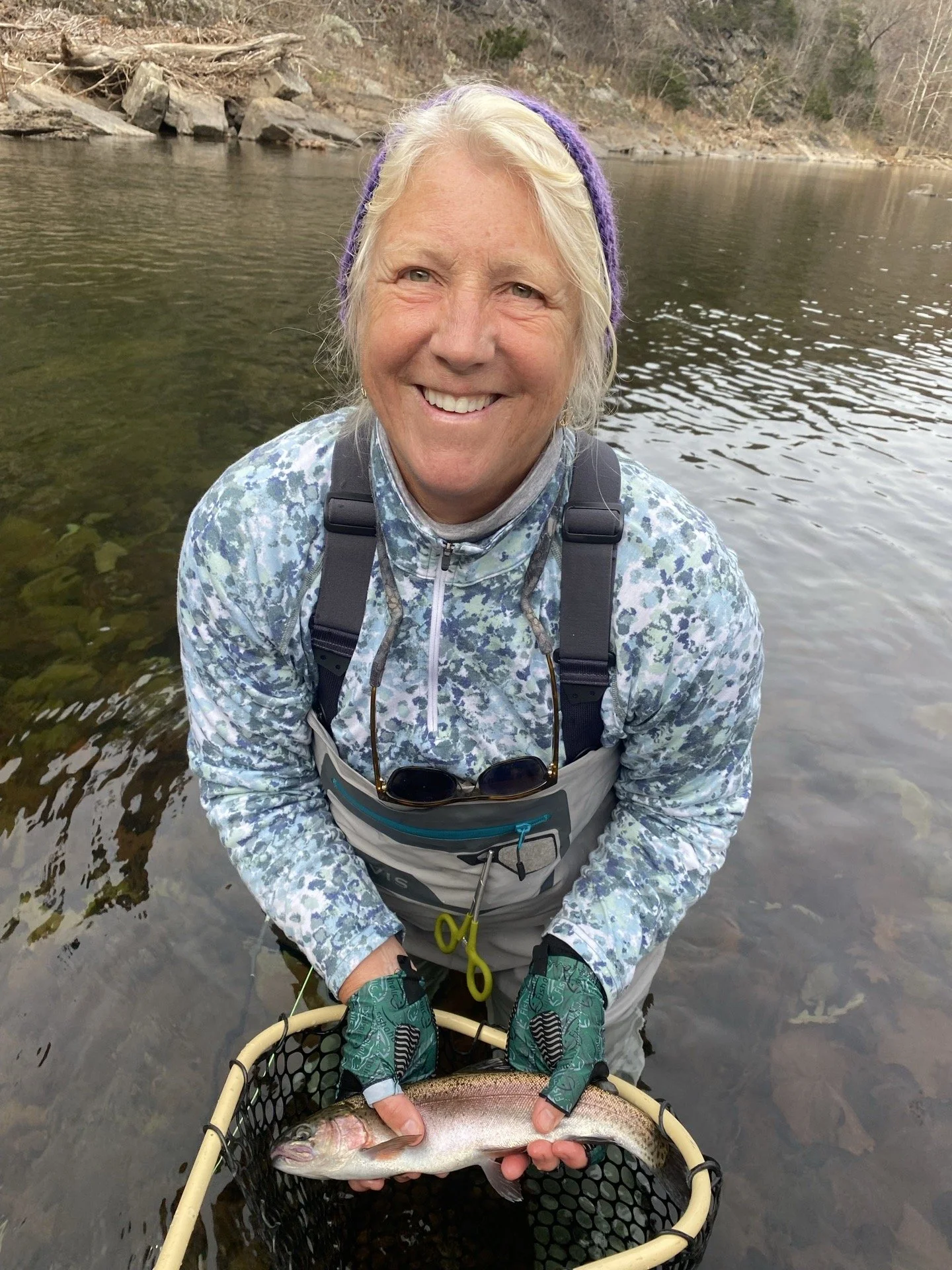 A woman in outdoor gear holding a fish over water, smiling at the camera.