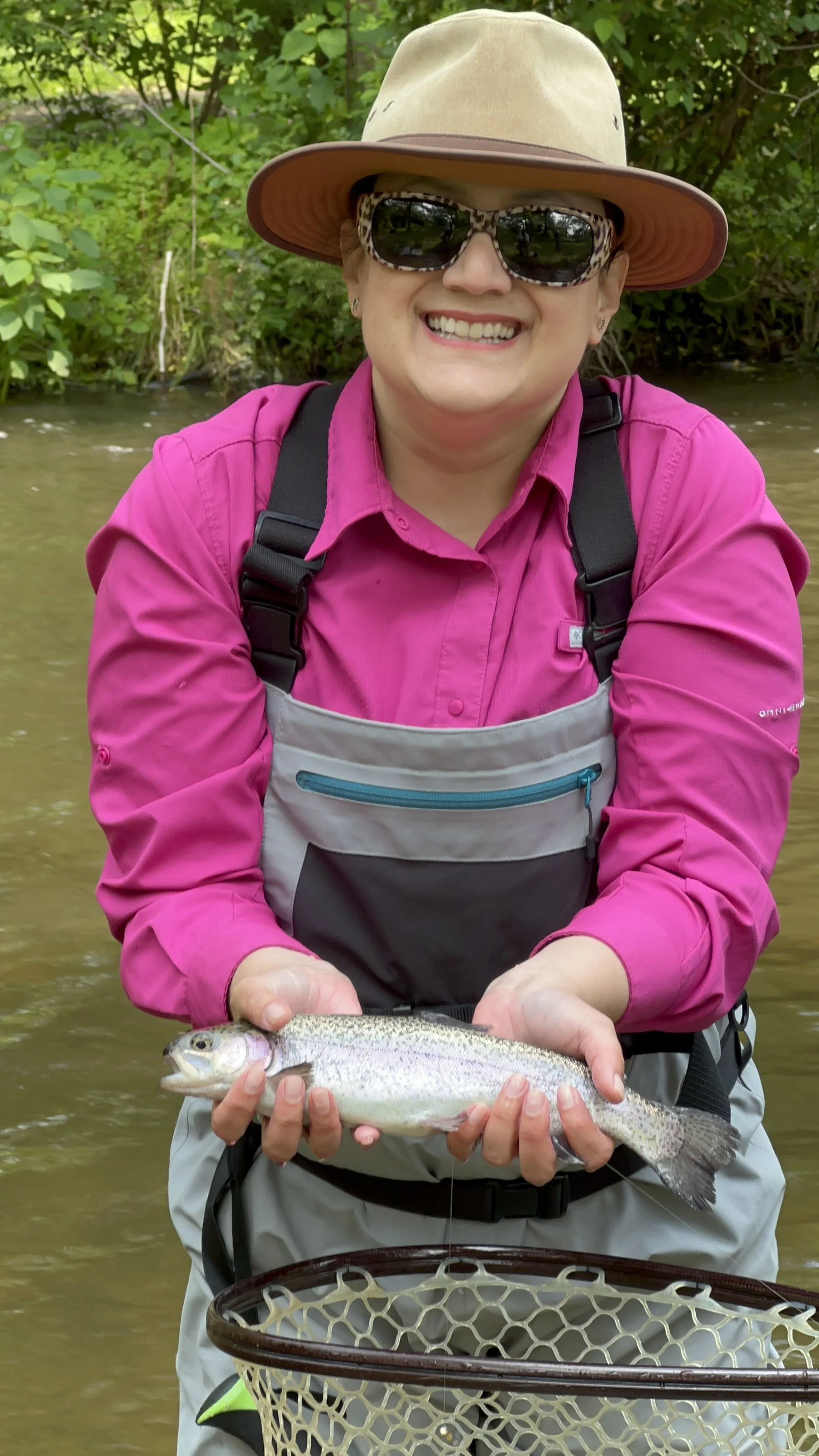 Person holding a small fish near a lake with leafless trees in the background.