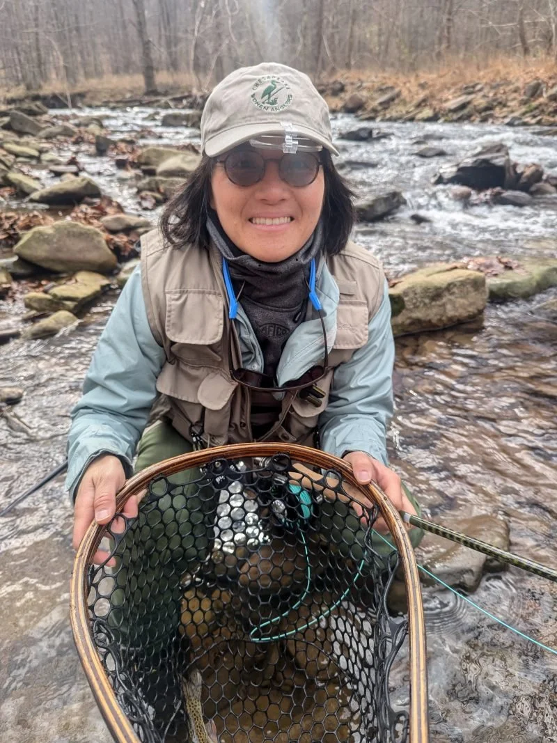 Woman wearing hat, sunglasses, and fishing gear holding a fishing net with a fish inside, standing in a rocky stream surrounded by trees.