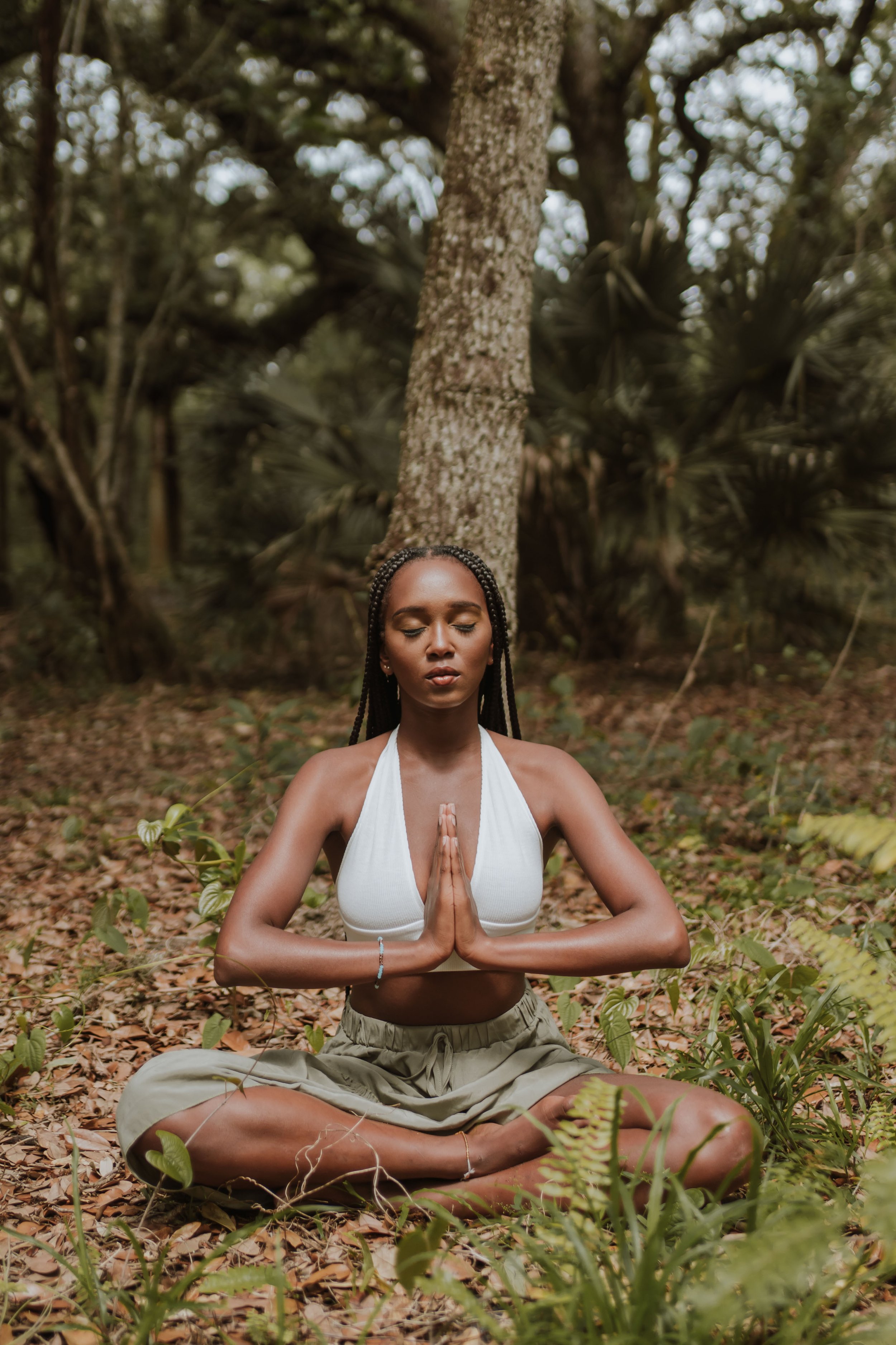 A woman practicing meditation outdoors in a forest, sitting cross-legged with hands in prayer position and eyes closed.