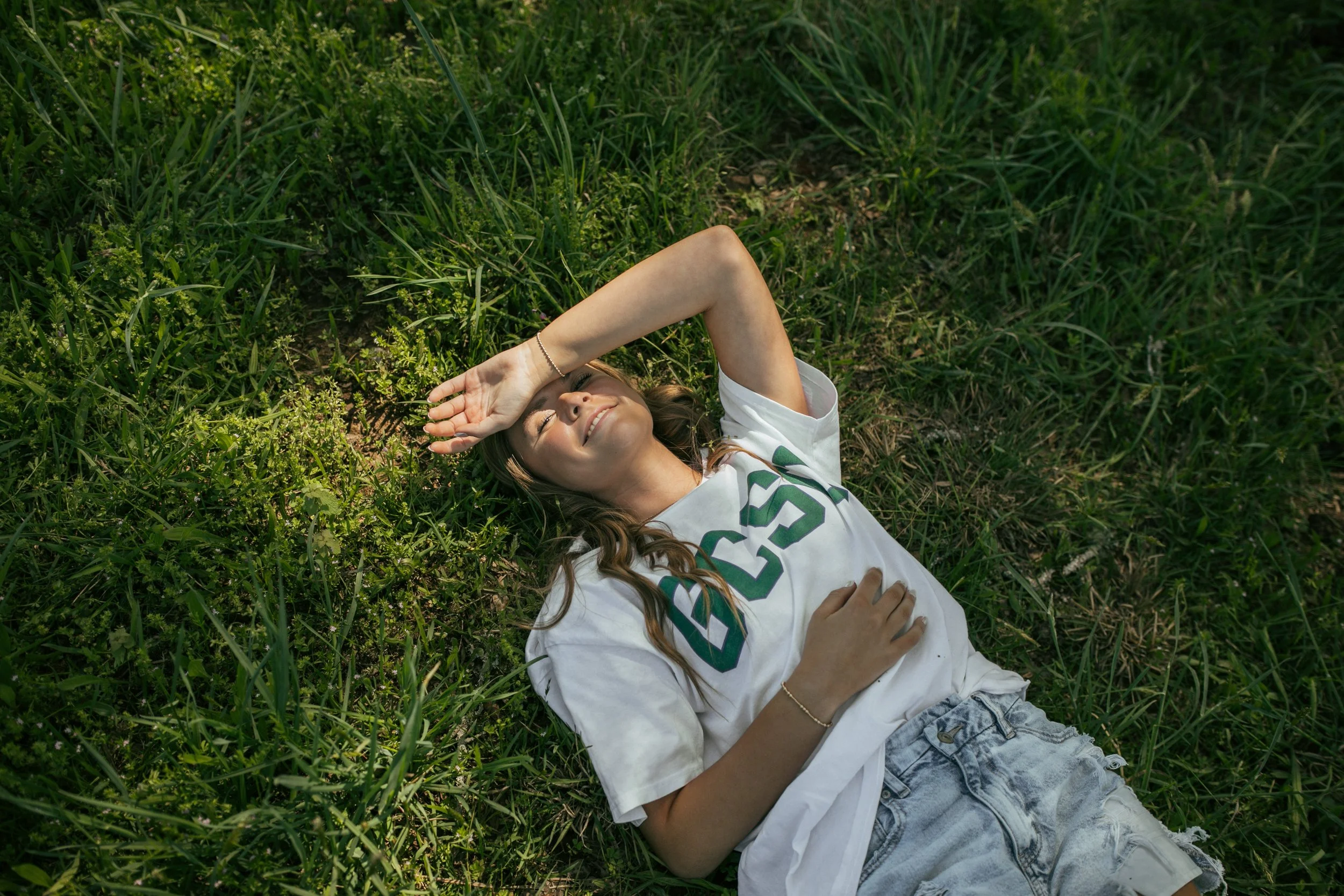 A young woman is lying on her back in a grassy area, smiling with one hand shielding her eyes from the sunlight and the other hand resting on her stomach.