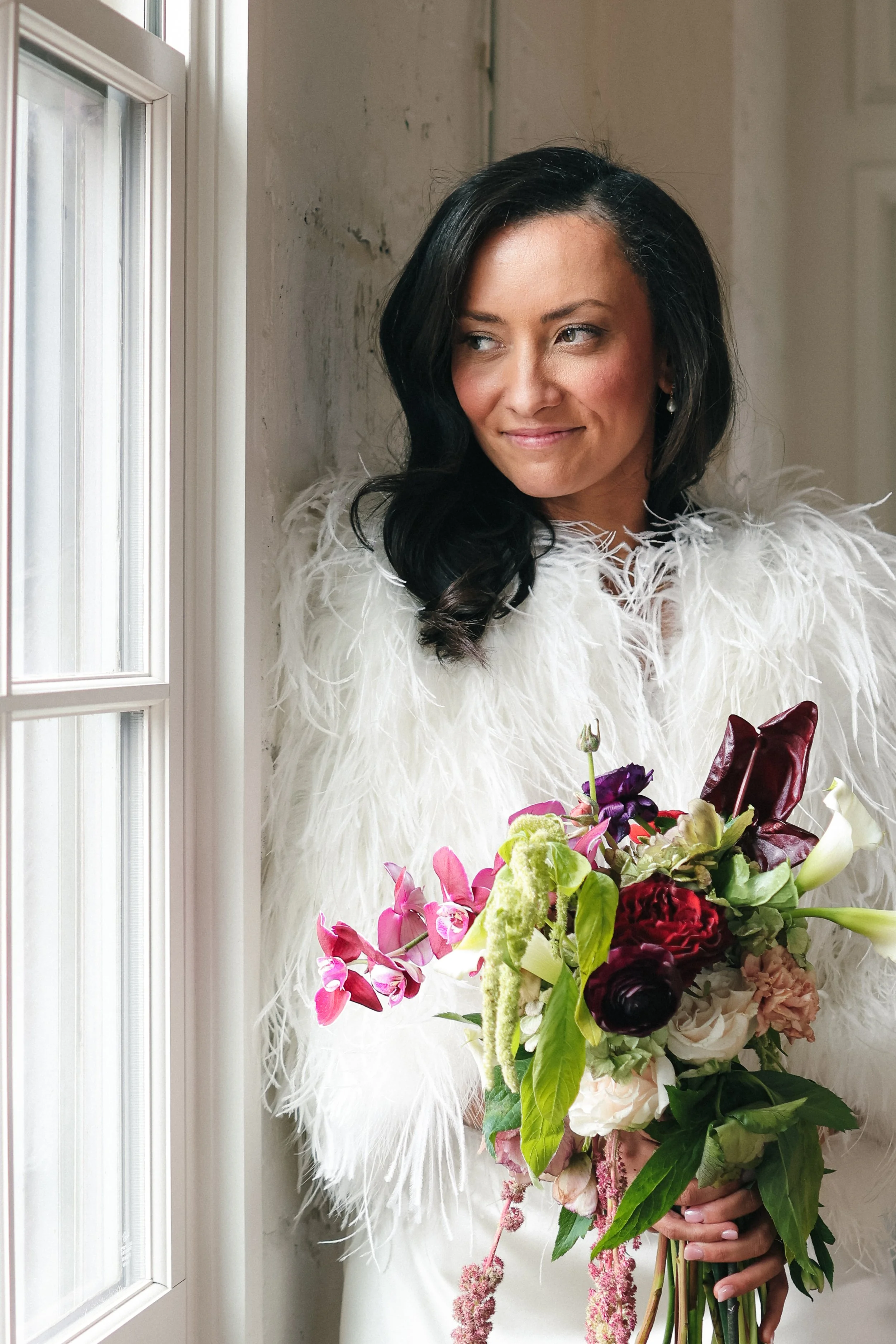 A woman in a white feathered dress holding a colorful bouquet of flowers next to a window.