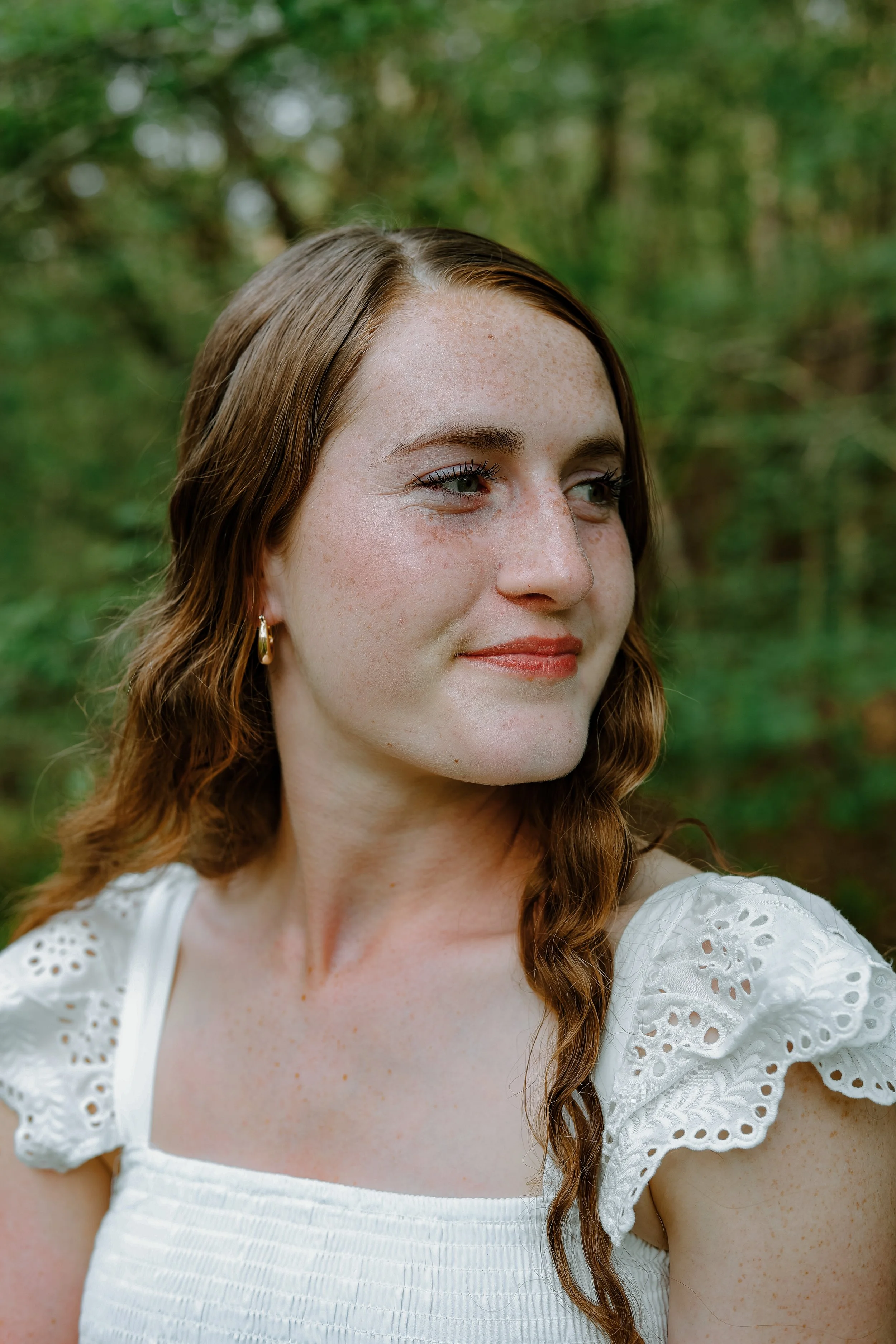Close-up of a young woman with reddish-brown hair and green eyes, wearing a white eyelet dress with puffed sleeves, outdoors with green foliage in the background.