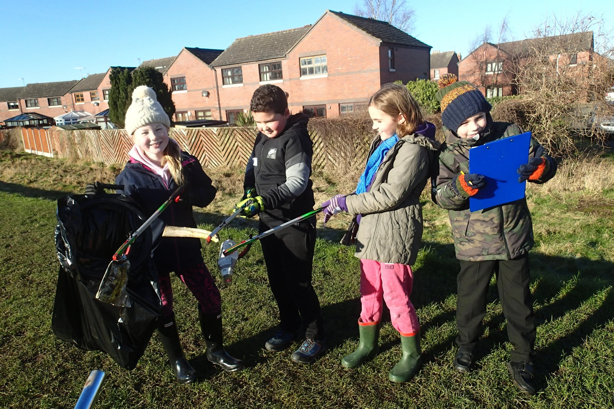 Four children outdoors, participating in a neighborhood cleanup, holding litter pickers and collecting trash, dressed warmly in jackets, hats, and gloves.