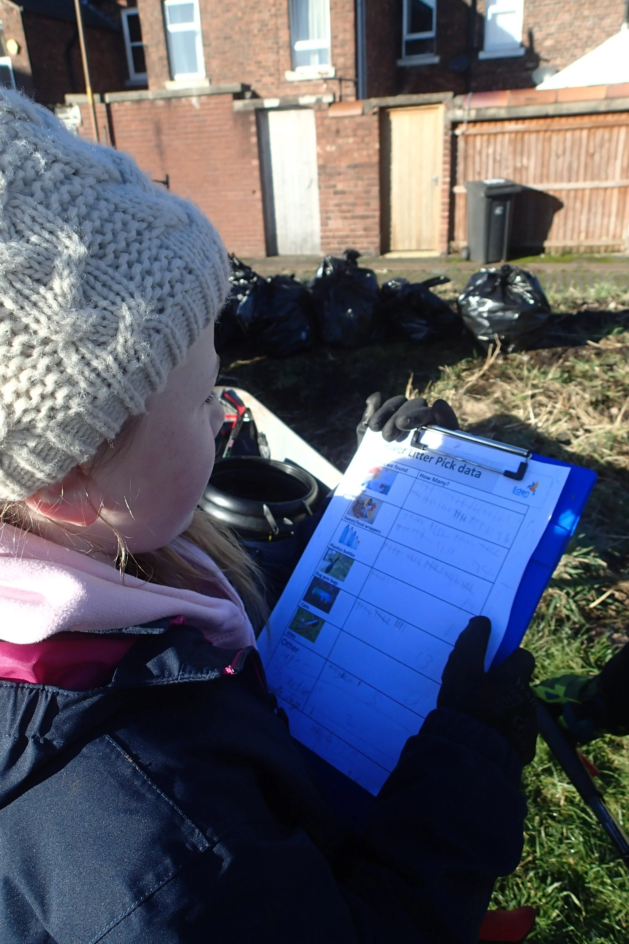 A young girl wearing a gray knit hat, pink hoodie, and black gloves holding a clipboard with litter pick-up data, outdoors on a sunny day near a brick building and black trash bags.
