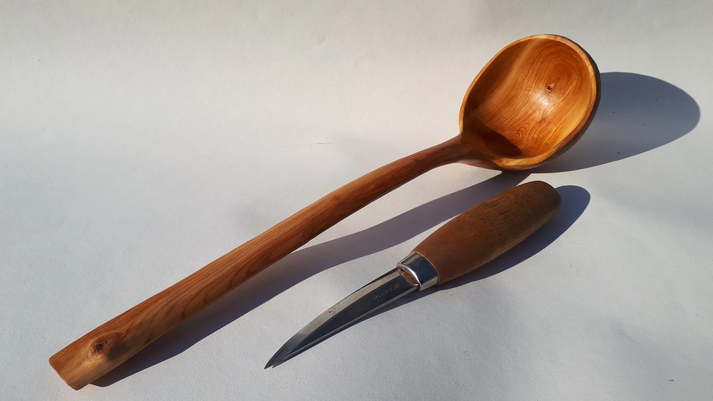 A wooden ladle with a long handle and a rounded bowl and a wood-handled knife with a curved blade lying on a white surface.