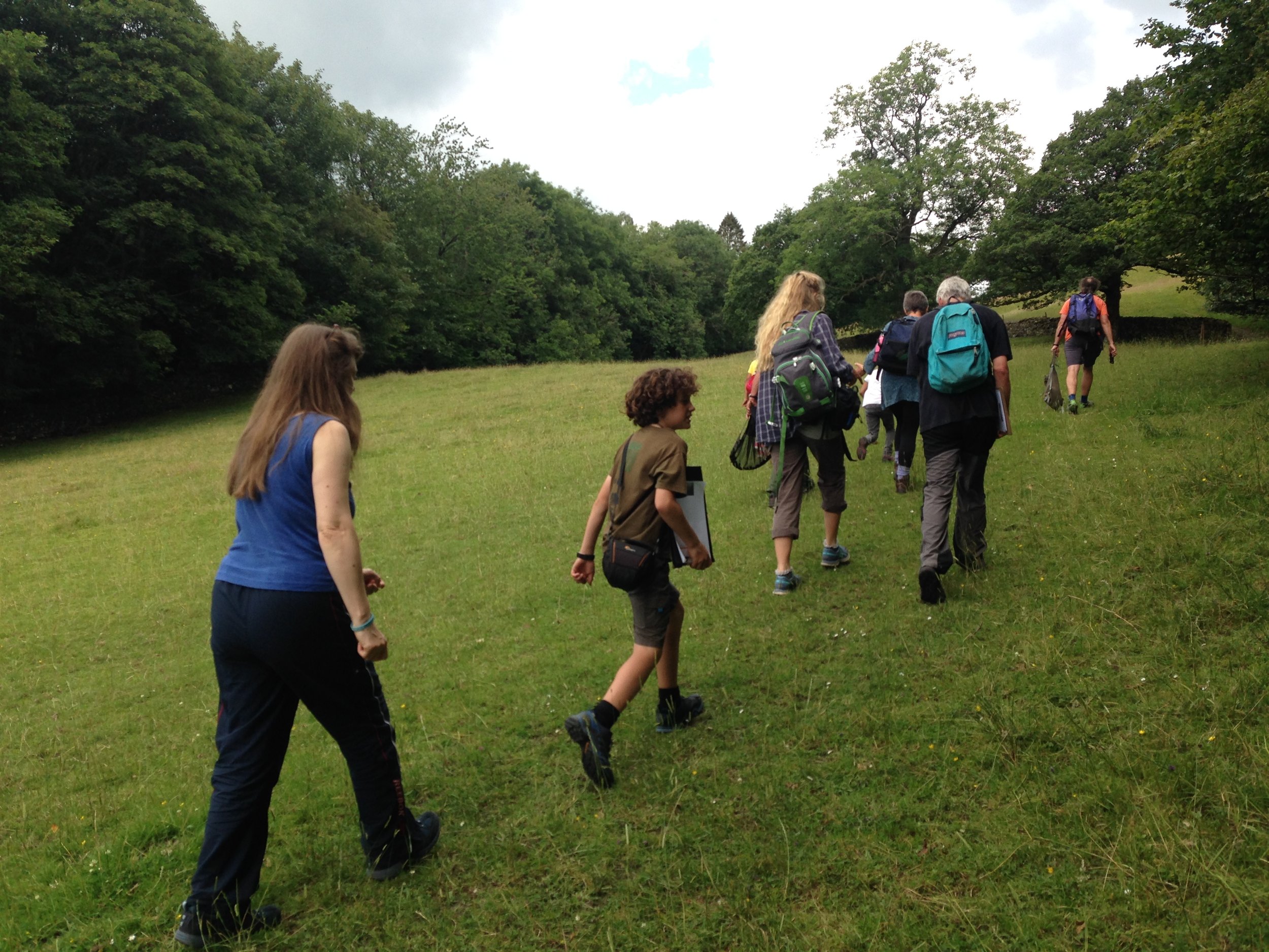 A group of hikers, including children and adults, walking uphill on a grassy trail in a forested area.