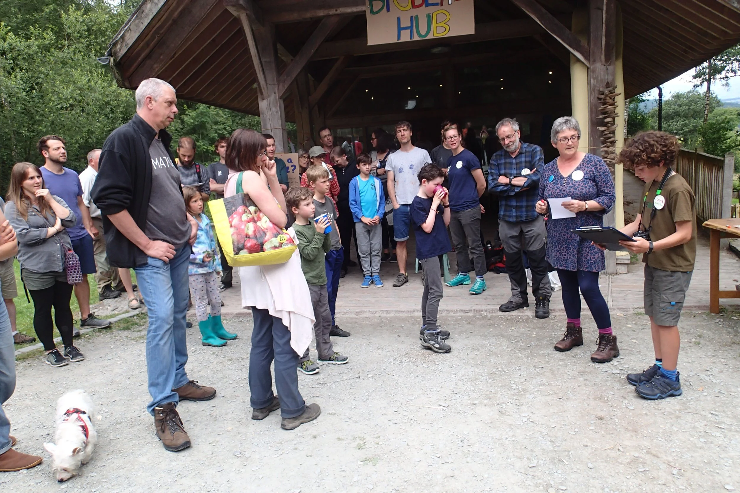 A group of people, including adults and children, gather outside a wooden building with a sign that reads "Bioblitz Hub." Some individuals appear to be listening while a woman with a clipboard and a young person with a camera are leading the session.