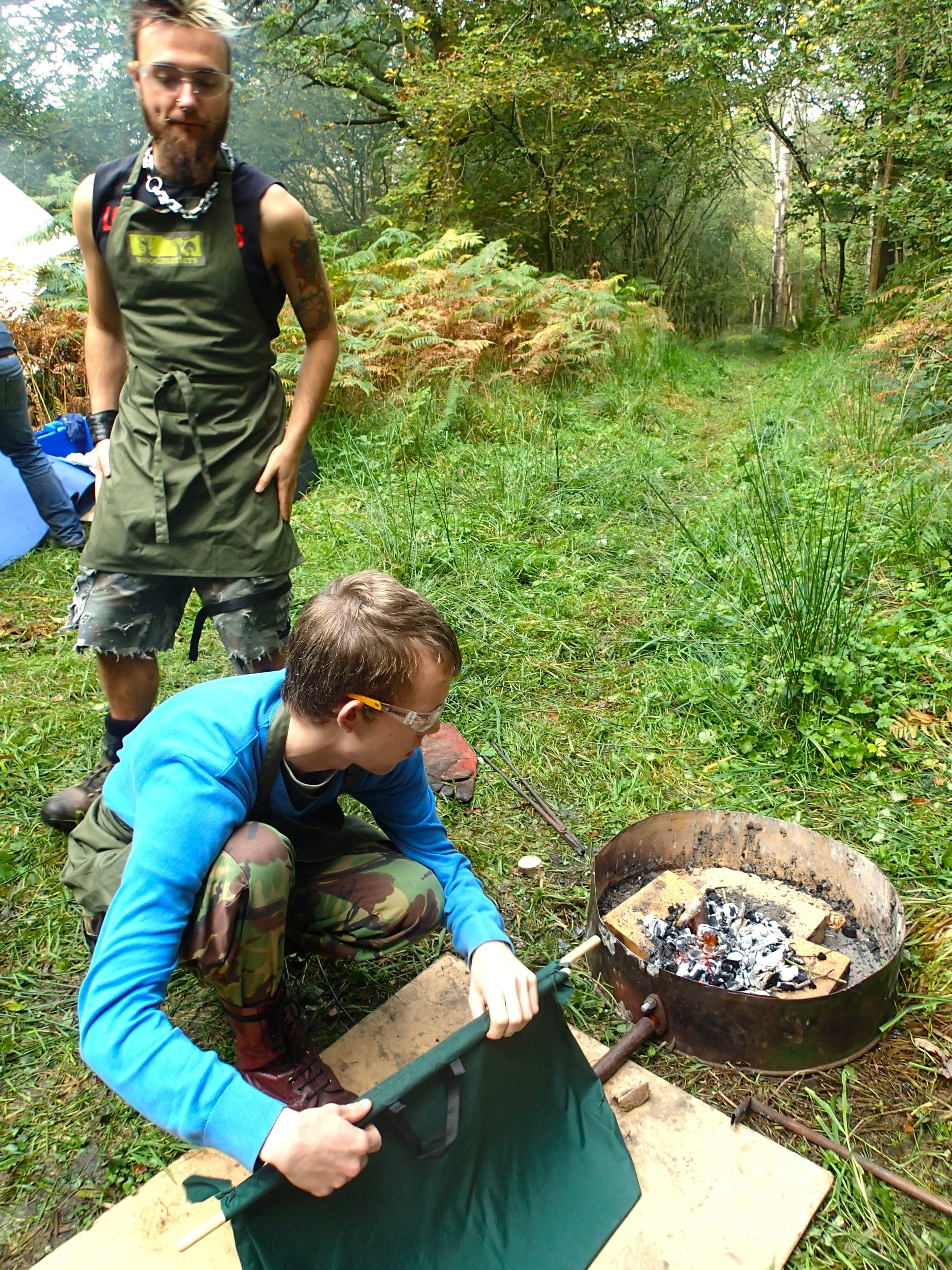 A person crouching by a portable outdoor fire pit with ash and charcoal, wearing safety glasses and camouflage pants, holding a green fireproof pad. Another person stands nearby in a green apron and casual clothing, observing in a forested outdoor se
