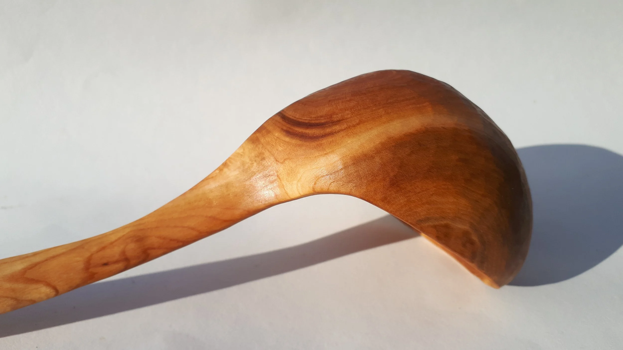 A wooden spoon with a rounded bowl and a long, smooth handle, casting a shadow on a light-colored surface.
