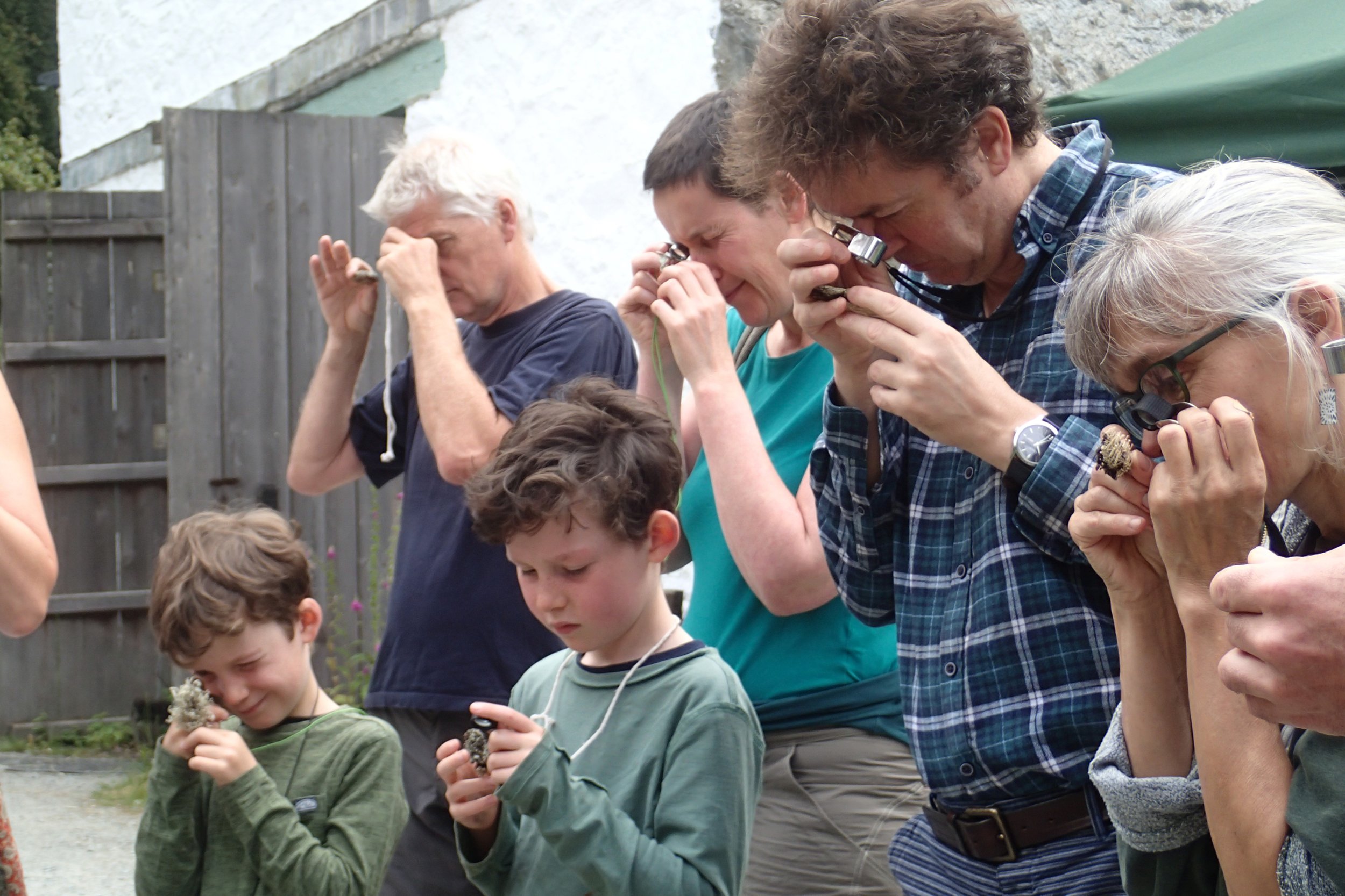 Group of children and adults closely examining objects with magnifying glasses outdoors.