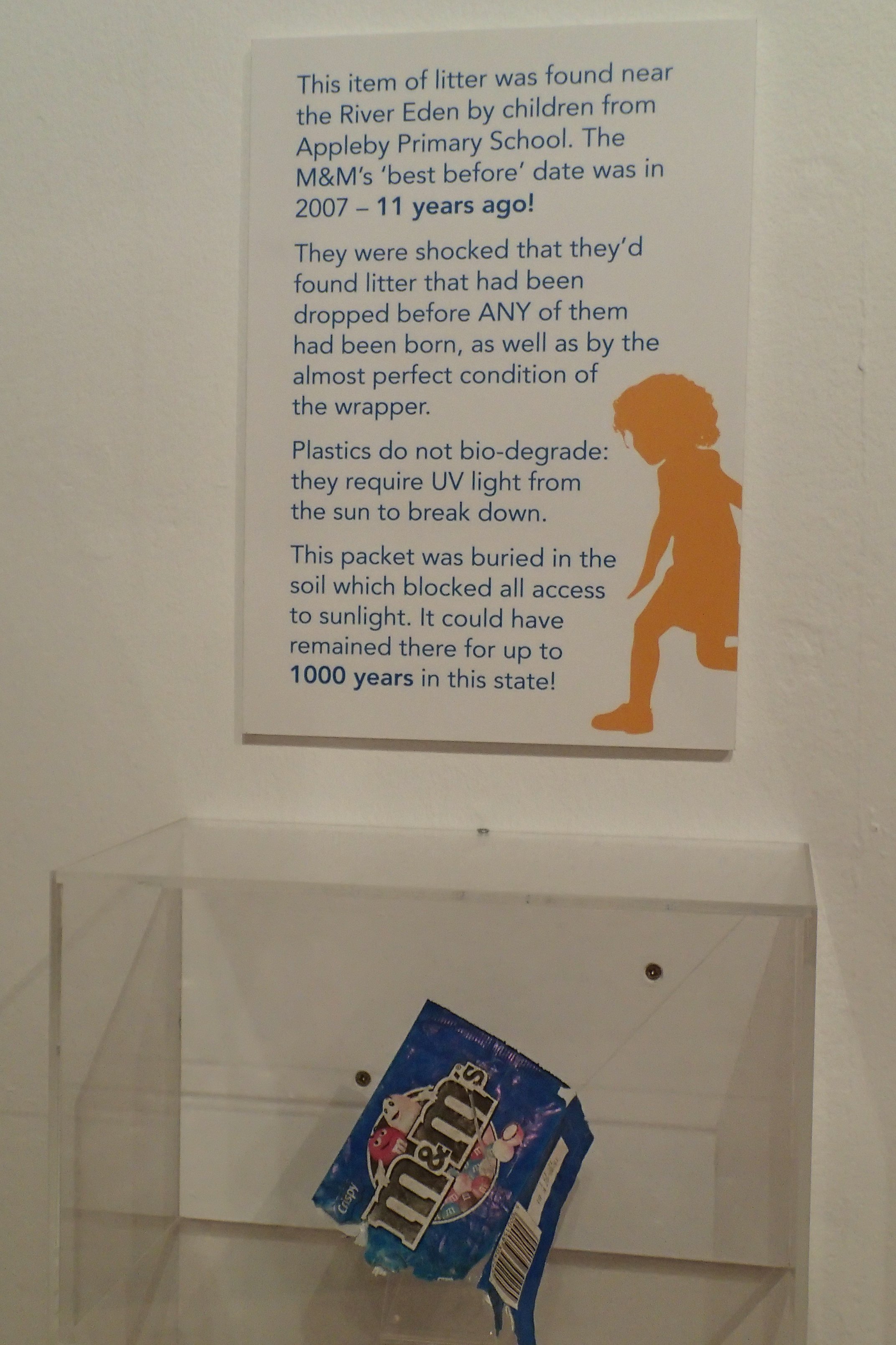 A plastic recycling or disposal box is empty except for a crumpled bag of M&M's candy inside. Above the box, a sign explains the significance of a piece of litter found near the River Eden, discovered by children from Appleby Primary School 11 years 