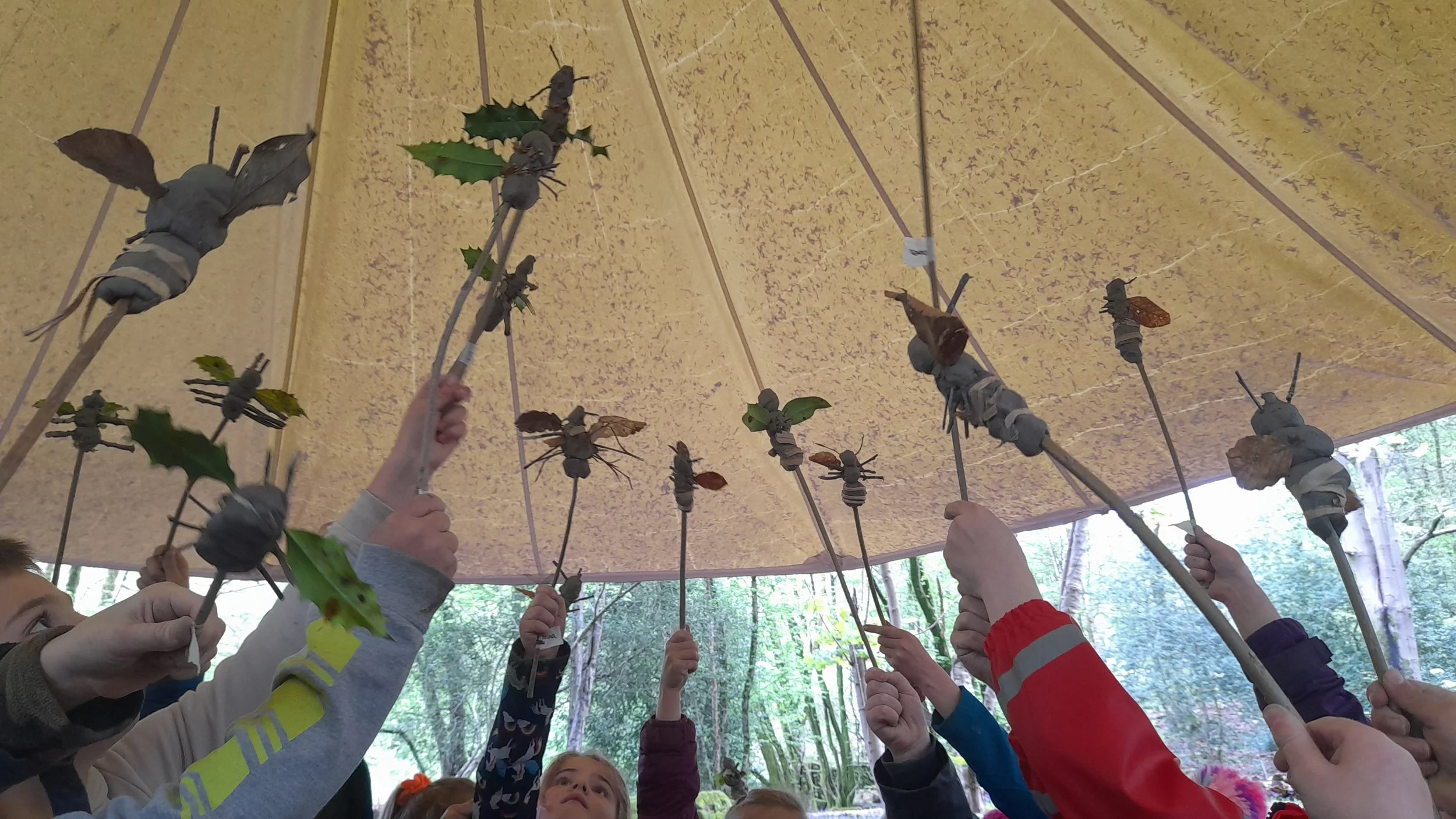 Children holding sticks with crafted insects, up towards a brown tent ceiling outdoors.