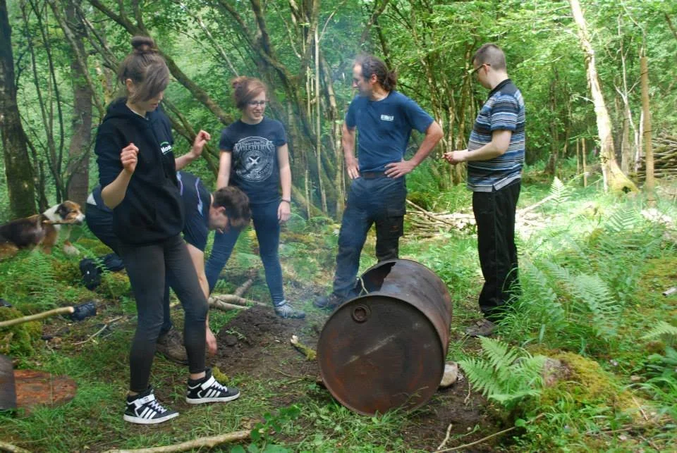 Group of people in a forest, gathered around a rusted barrel on the ground. Some are observing, one person is bending down, and a dog is nearby.