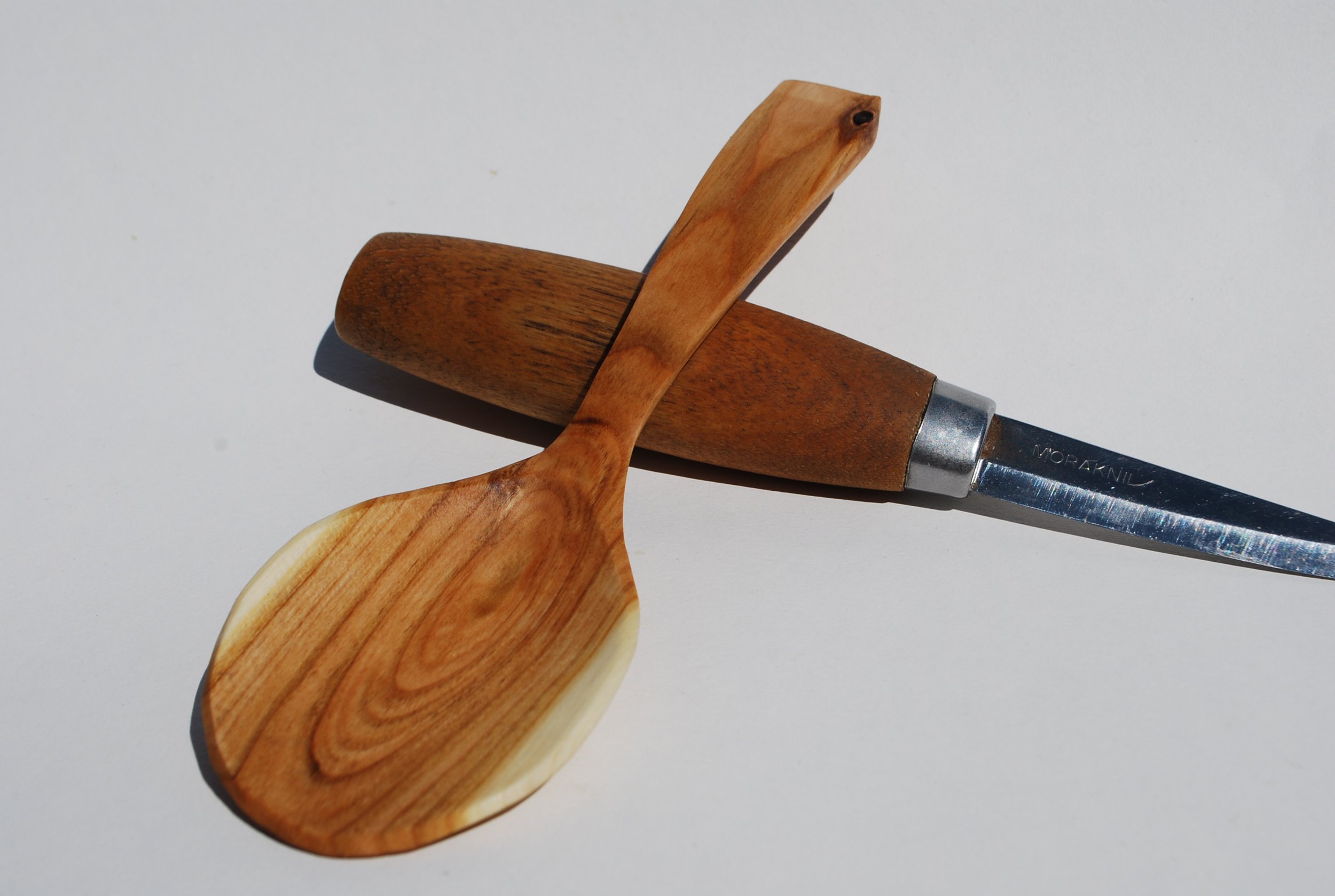 A wooden spatula with a natural wood finish resting on a wooden rolling pin, which is lying on a plain white surface.