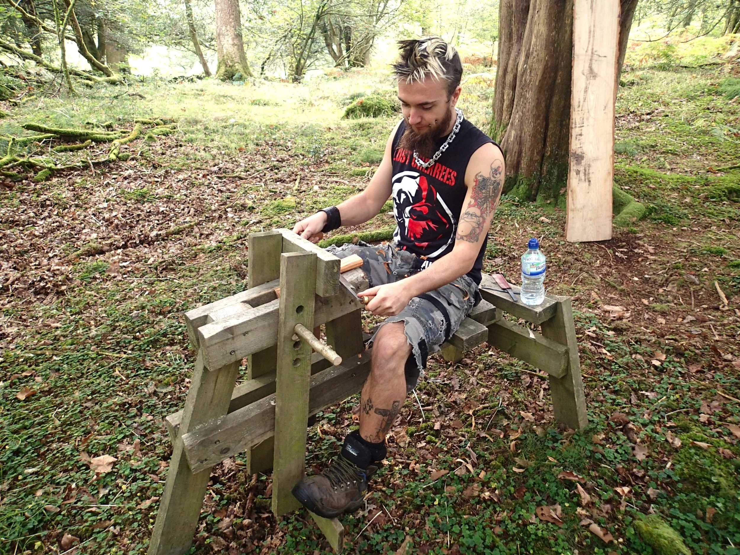 A young man with tattoos and a punk hairstyle working on a woodworking project outdoors in a forest. He is seated on a makeshift workbench and using a hand tool, with a water bottle beside him.