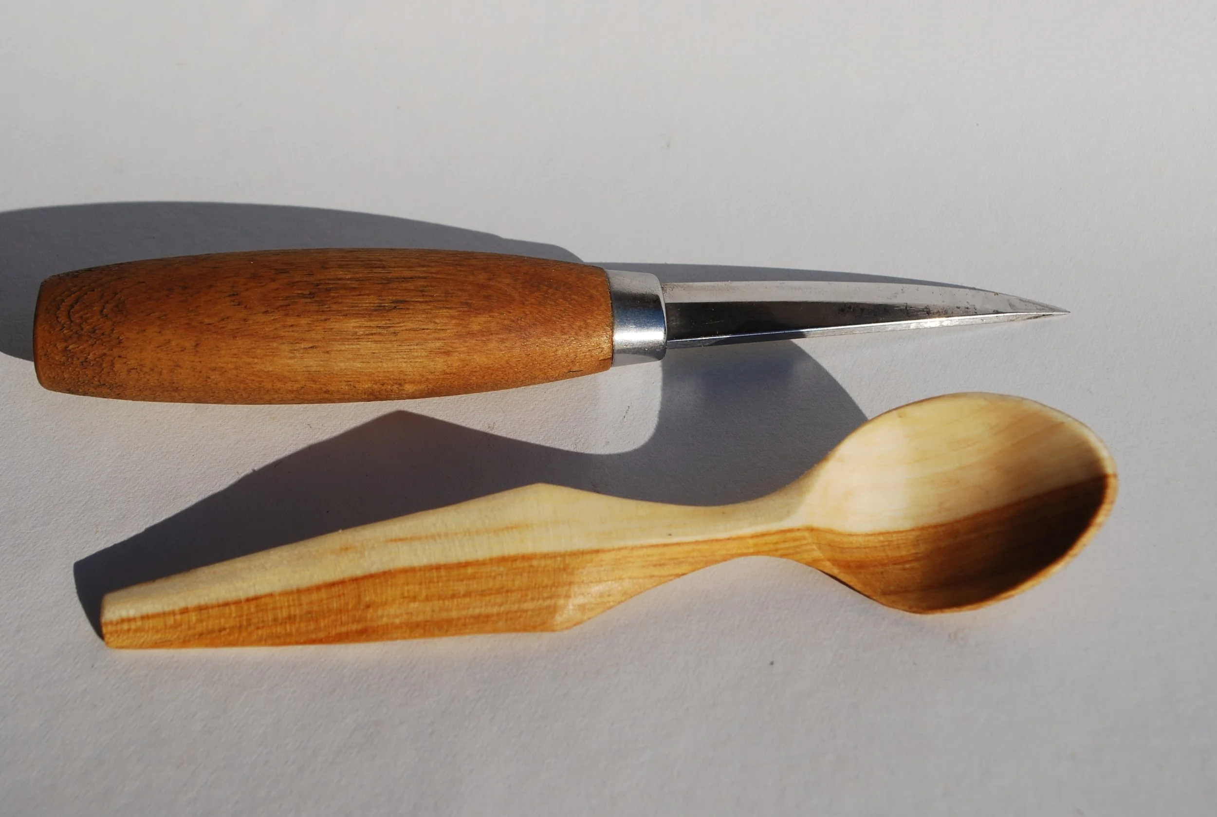 A kitchen knife with a wooden handle and a sharp metal blade aligned with a wooden spoon on a white surface.