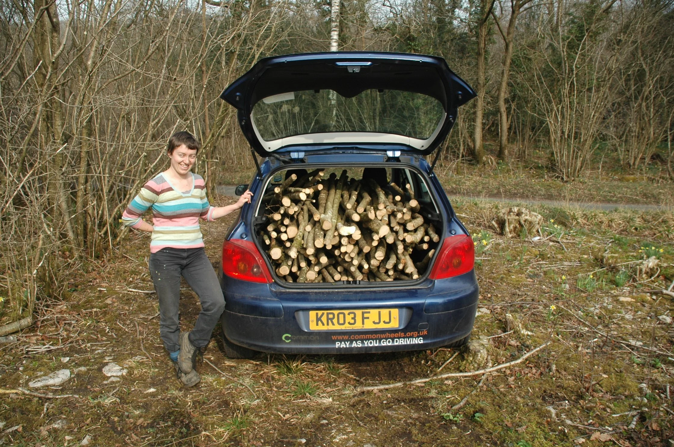 A woman standing next to the open trunk of a blue car filled with cut logs, in a wooded area.