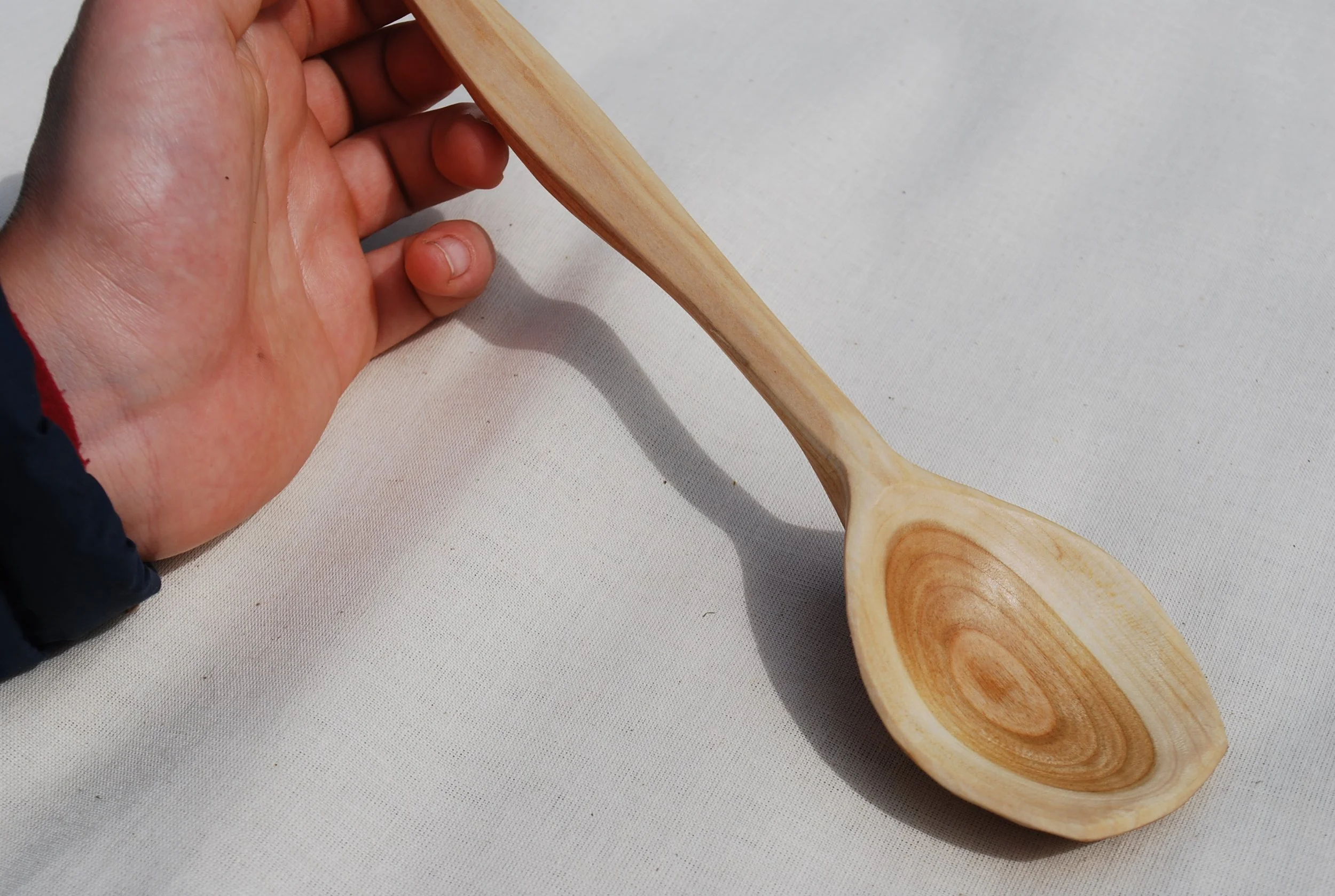 A hand holding a wooden spoon with a round, shallow bowl, sitting on a white fabric surface.