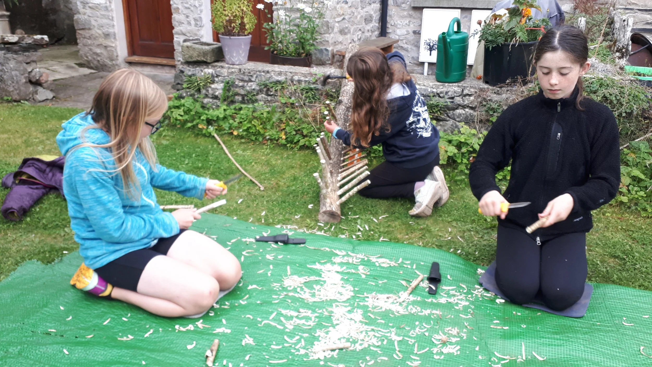 Three girls kneeling on a green mat outdoors, carving wood with small hand tools, with wood shavings scattered on the mat. One girl on the right in a black jacket is focused on her work, the girl in the middle in a dark hoodie is carving wood as well