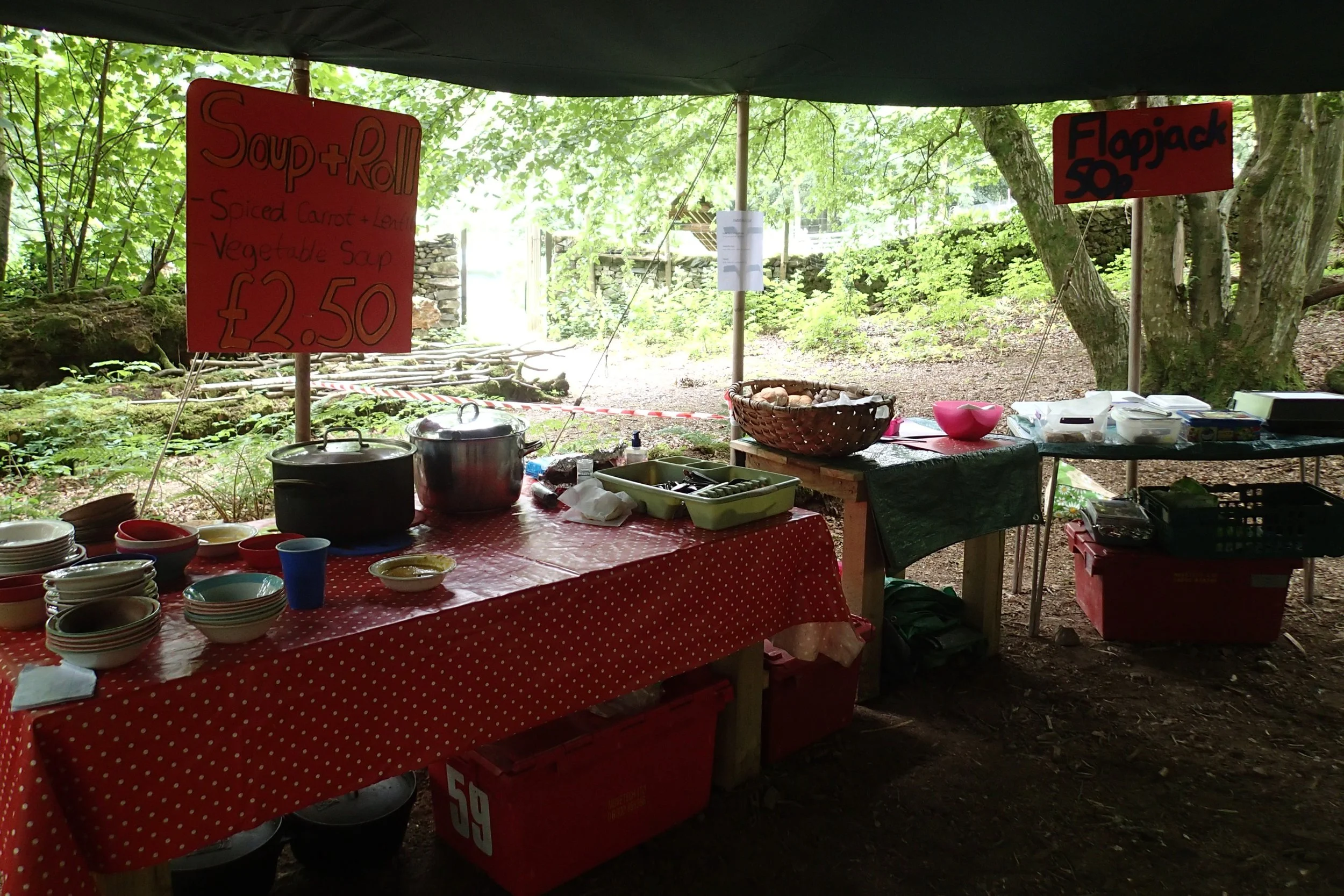 Outdoor food stall under a green canopy with signs advertising soup and roll, with various bowls, pots, and containers on tables, set in a wooded area.