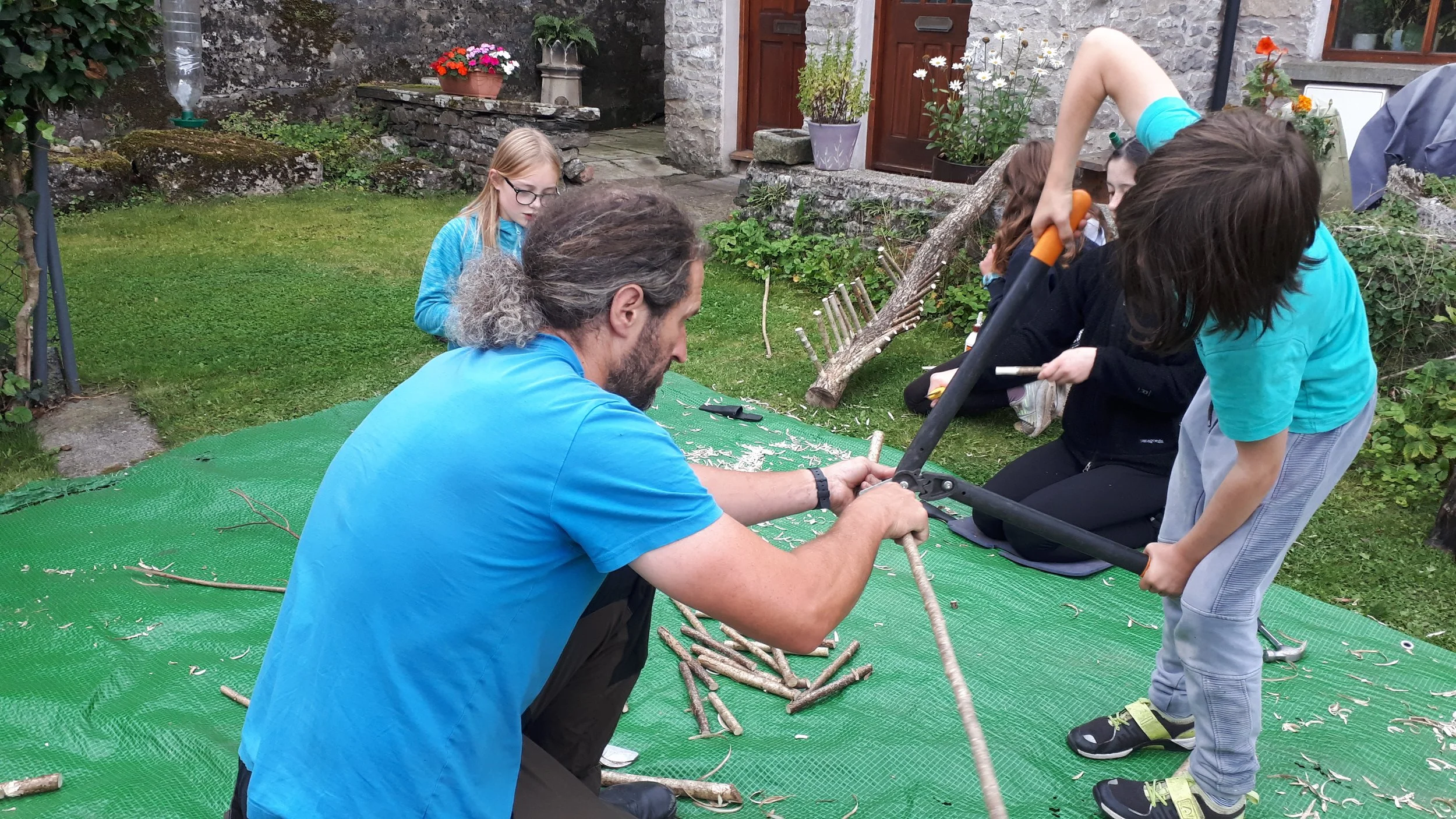 A man and a boy are sawing a long branch together with a two-person saw on a green tarp in a backyard. Two girls are sitting in the background, and another girl is standing nearby watching. The backyard has a stone wall, flowers, and garden tools.