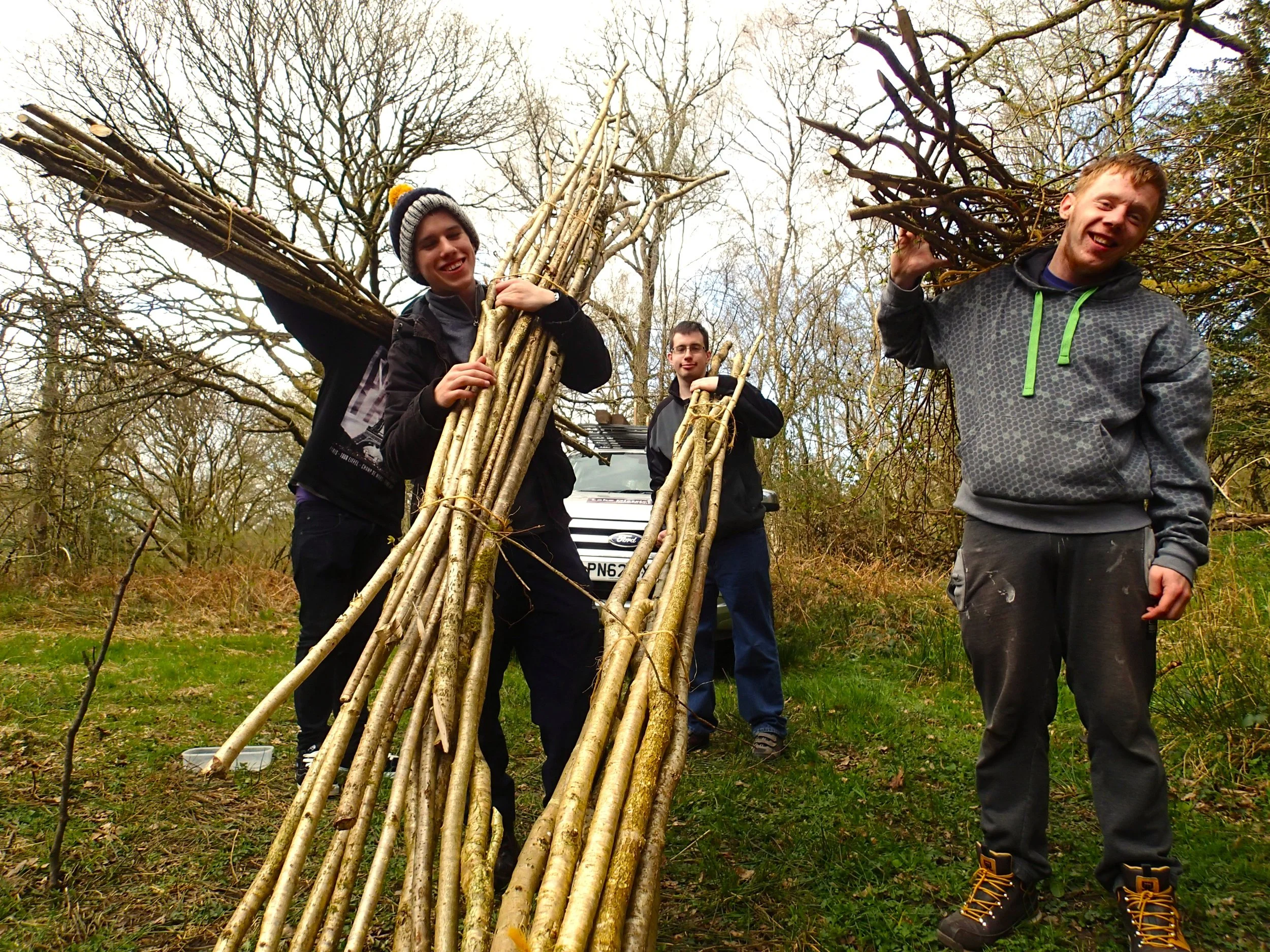 Four young men holding sticks and branches in a wooded outdoor area, smiling and posing for the camera.