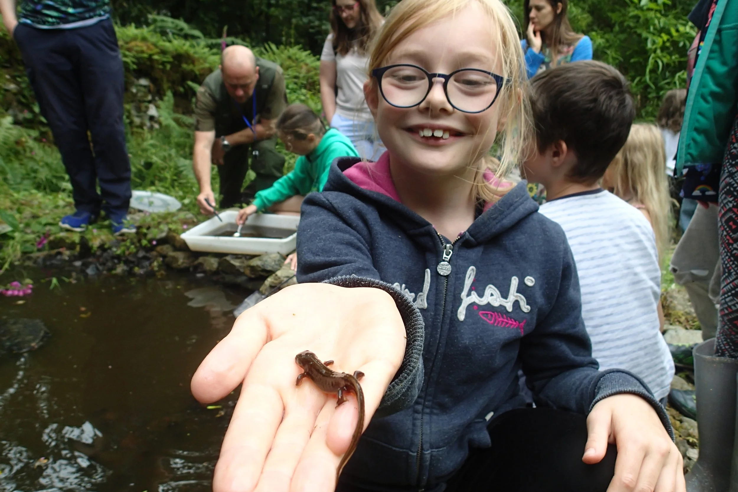 A young girl smiling and holding a small newt in her hand during a nature outing, with a group of children and adults exploring near a pond in a forest setting in the background.