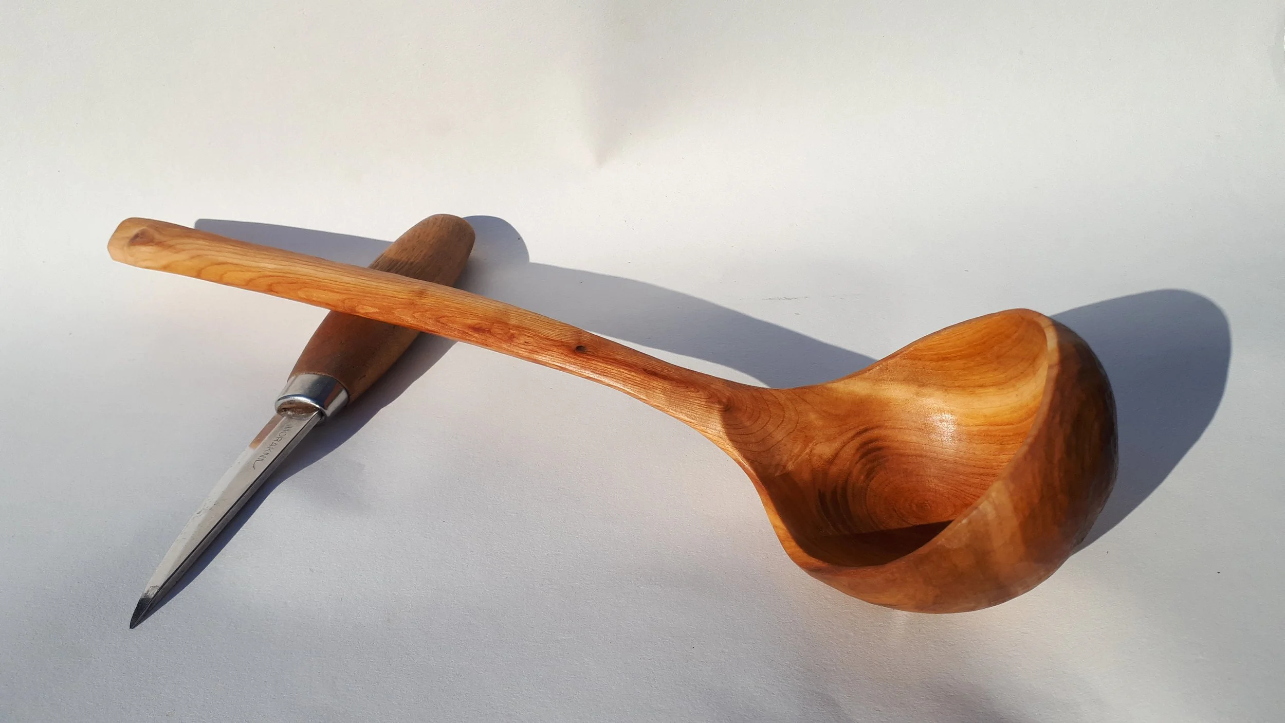 A wooden scoop, a wooden-handled knife, and a wooden spoon on a white surface with a shadow cast from sunlight.