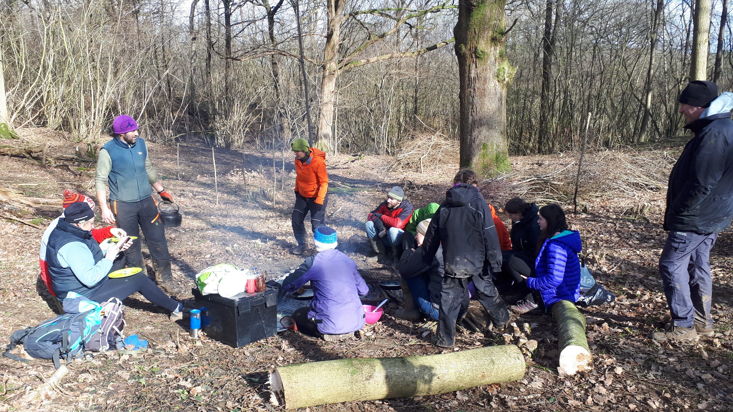 Group of people gathered outdoors in a wooded area, sitting and standing around a small campfire, with trees and fallen logs in the background, dressed in outdoor clothing for cool weather.