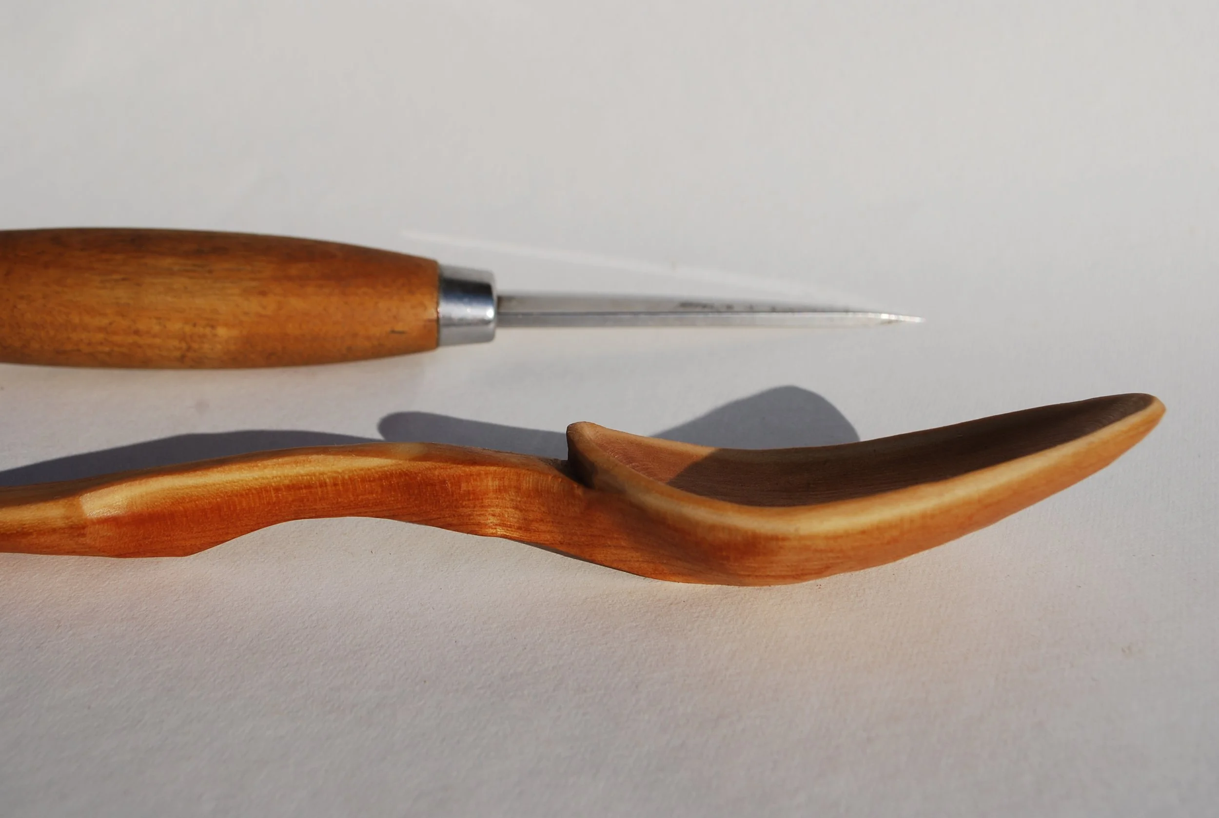 A wooden-handled craft knife and a wooden spoon lying on a white surface.
