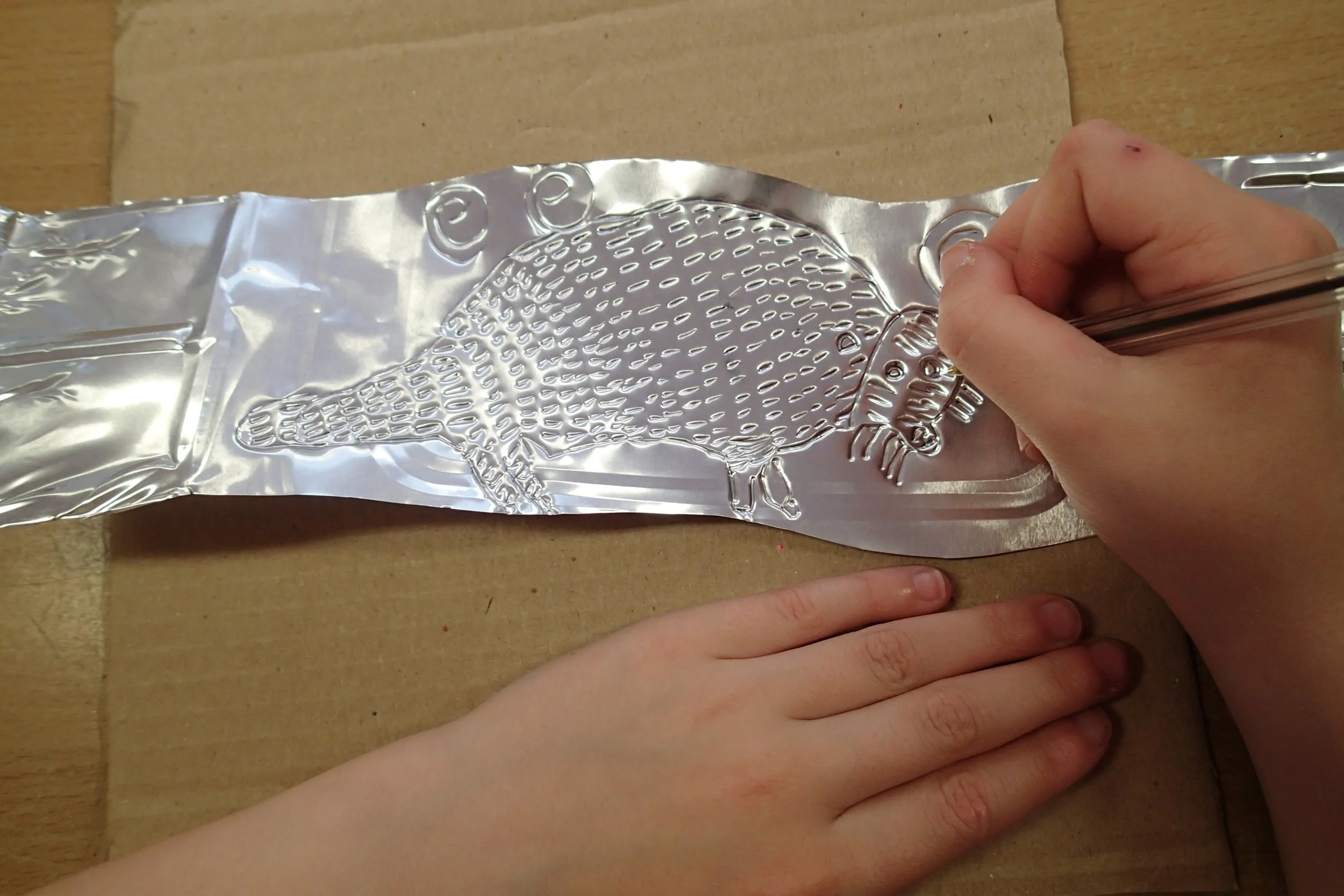 A person is embossing an image of an armadillo onto a piece of aluminum foil with a tool, while a child's hand rests nearby on a wooden surface.