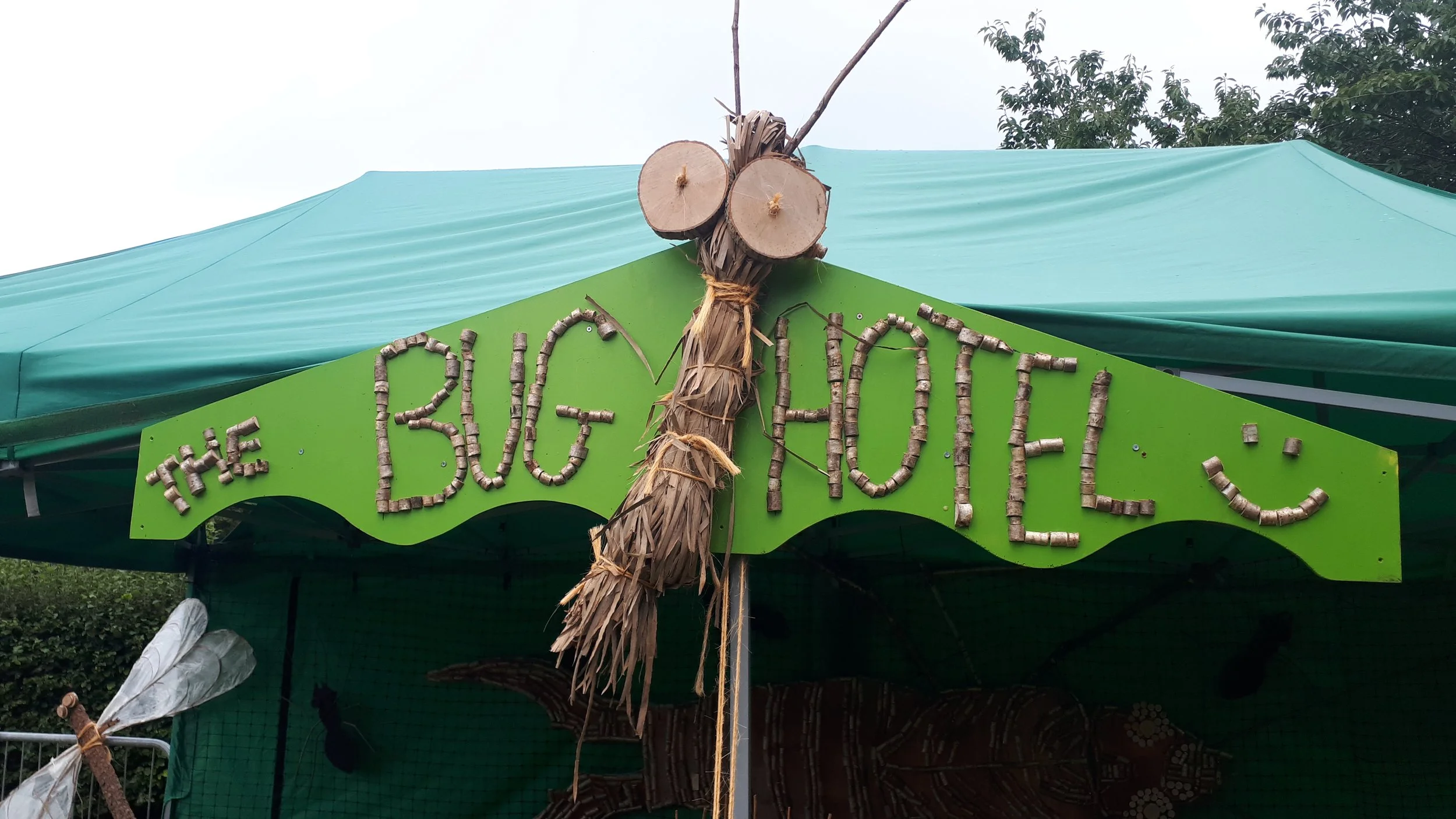 A sign that reads 'The Bug Hotel' made from small wooden sticks, with a large bug figure constructed from sticks and two round wood slices for eyes, hanging from a green tent canopy.