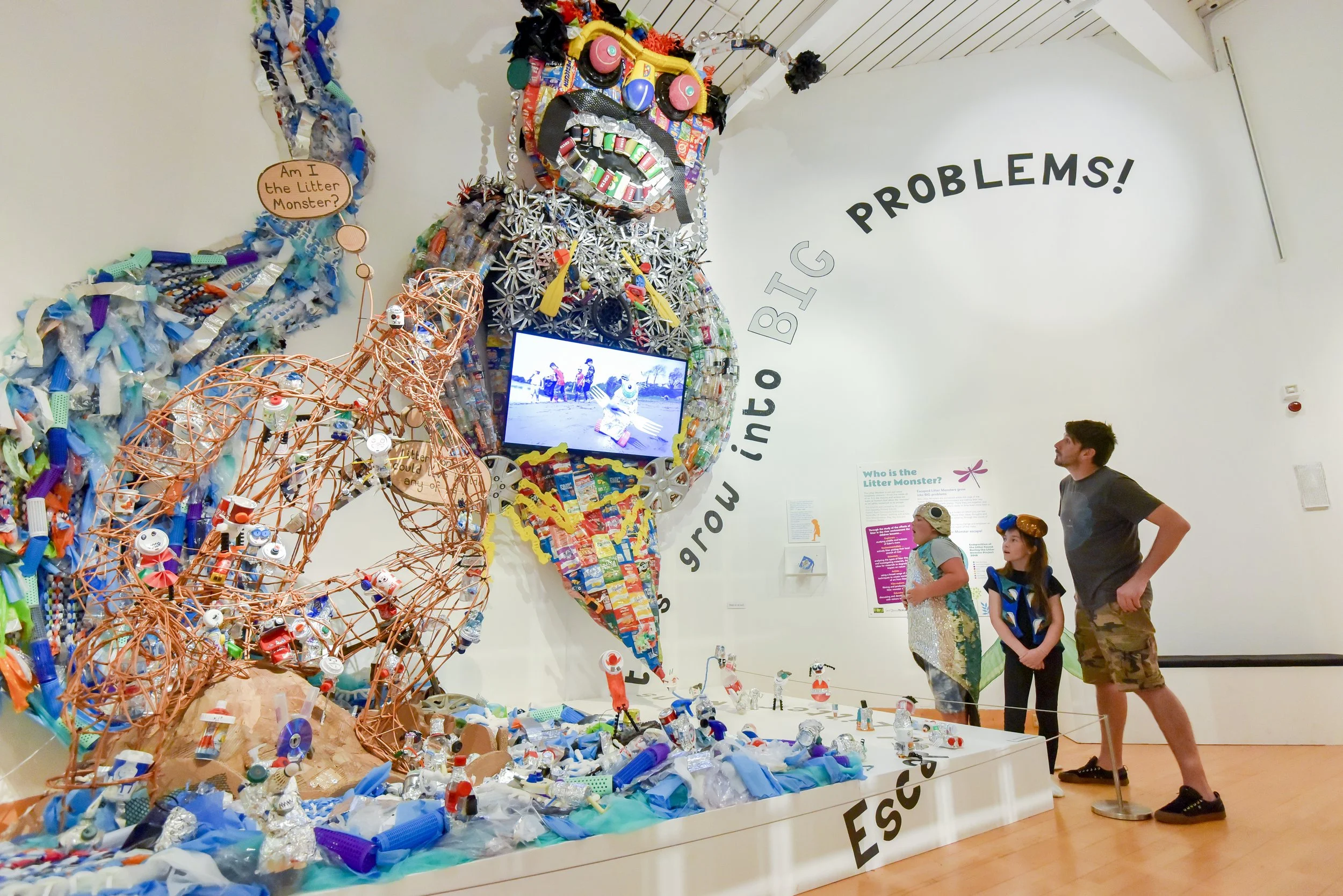 A large colorful sculpture of a monster made from recycled materials, including bottle caps, cans, and plastic bottles, displayed in an art exhibit with three visitors observing it.