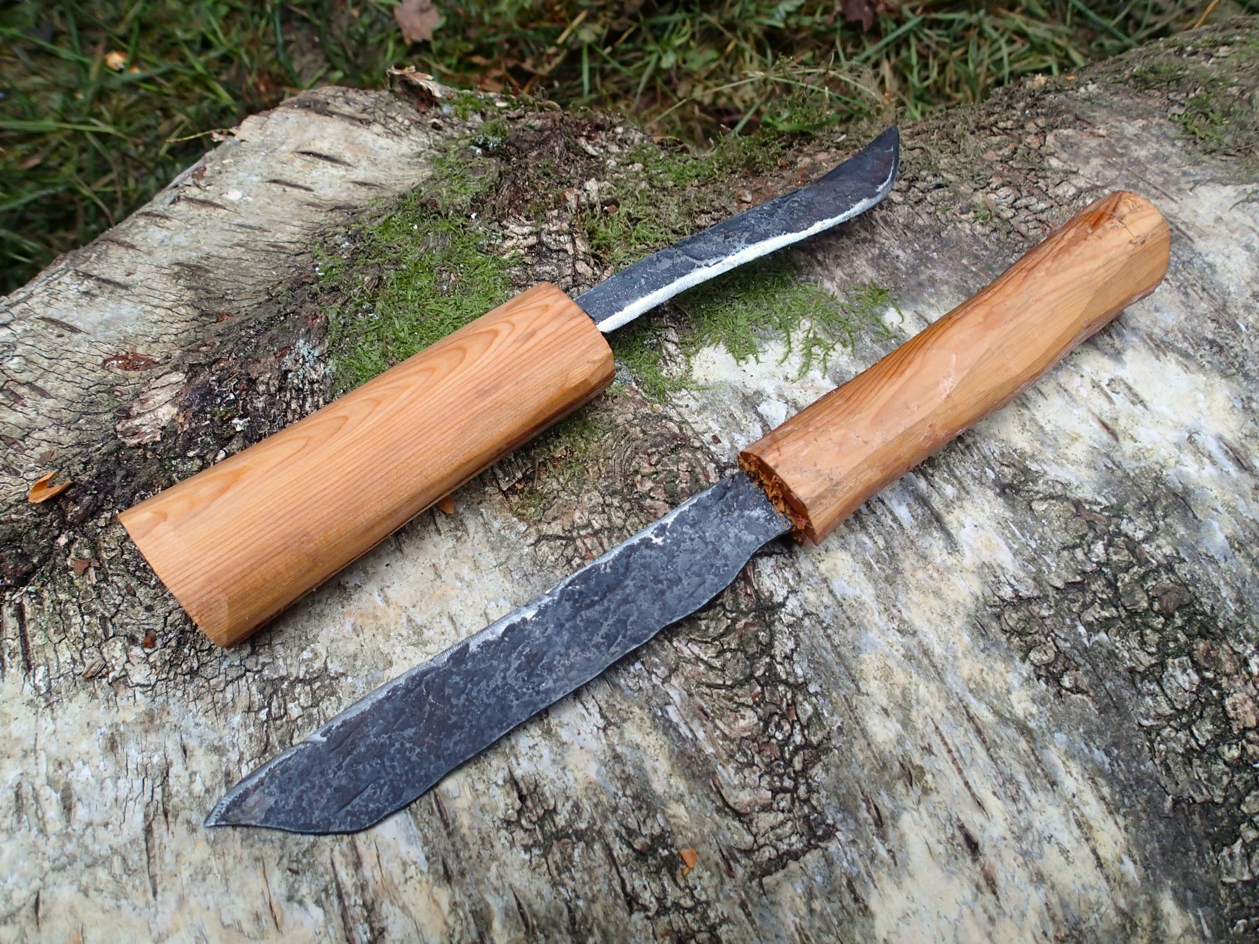 Two handcrafted knives with wooden handles resting on a moss-covered log outdoors.