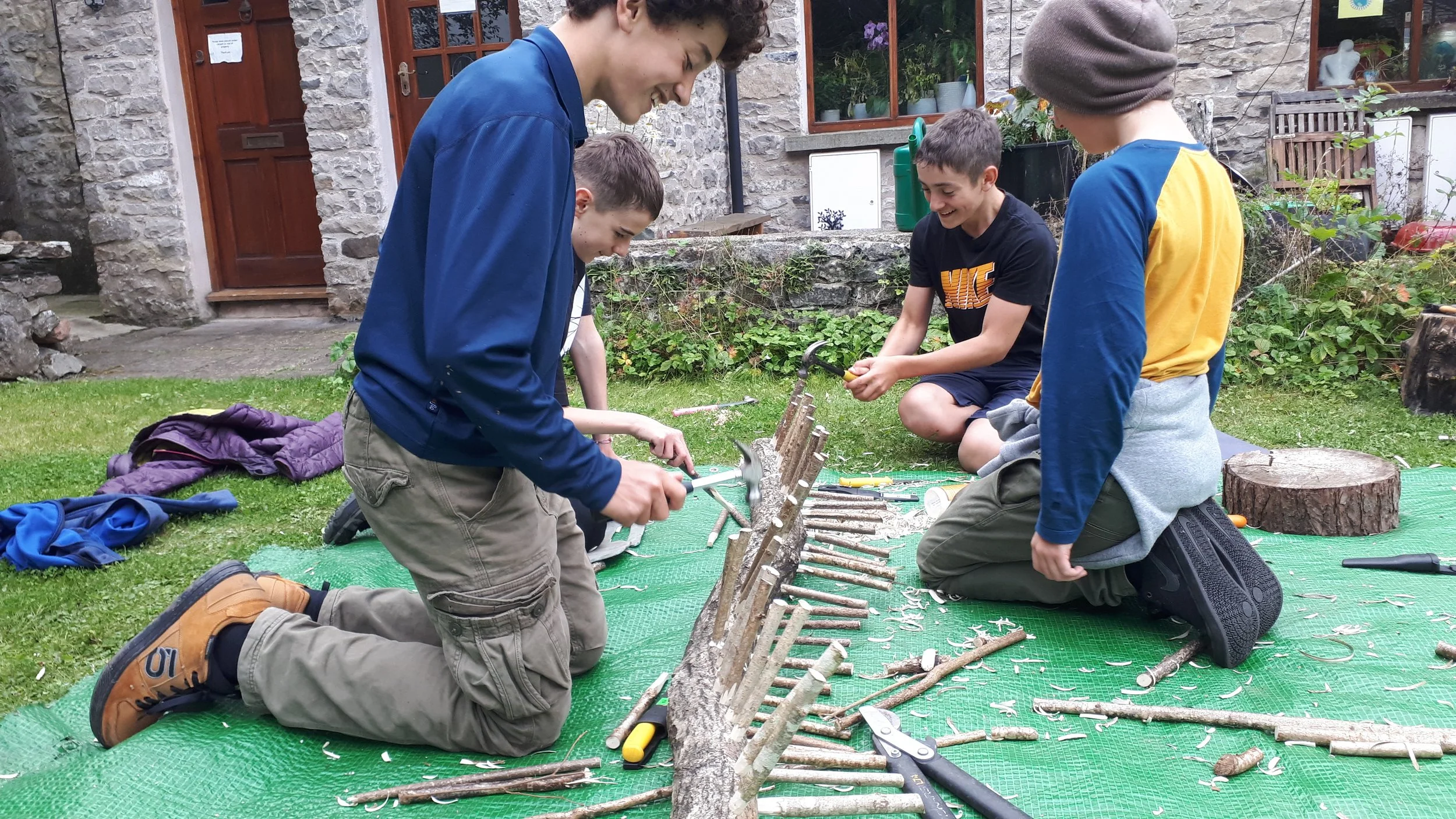 Group of children kneeling on a green mat outdoors, using tools to carve sticks and branches, with a stone house and garden in the background, engaged in crafting or woodworking activity.