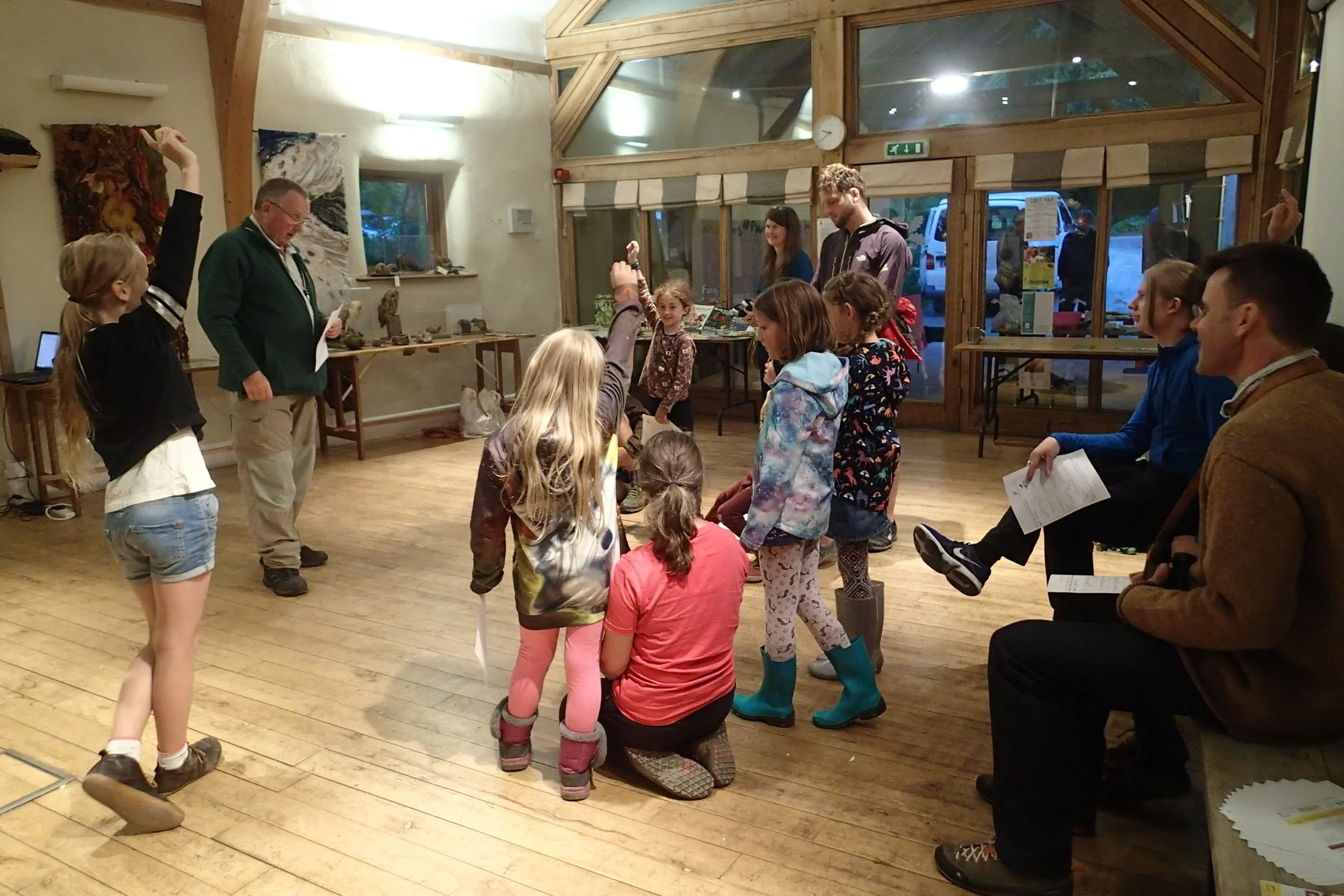 Children and adults gathered in a room, with some children raising their hands and others sitting or kneeling on the wooden floor. Adults are seated or standing nearby, some holding papers, and some children are wearing colorful jackets and boots. Th