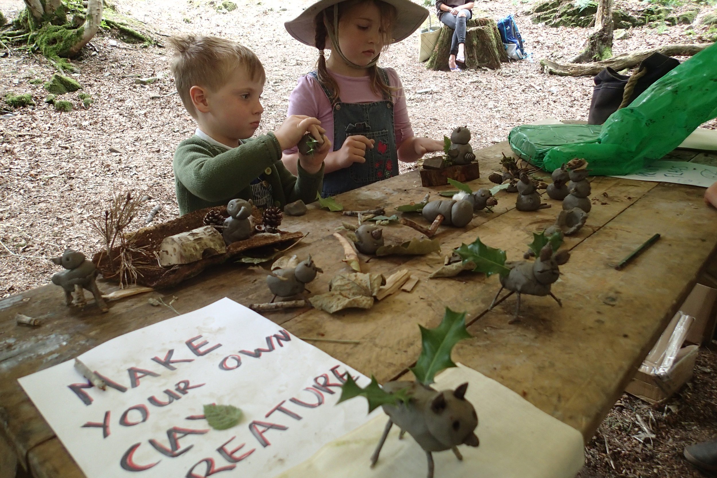 Two children, a boy and a girl, are making craft figures out of clay and natural materials like leaves and pinecones on a wooden table outdoors in a forest setting.