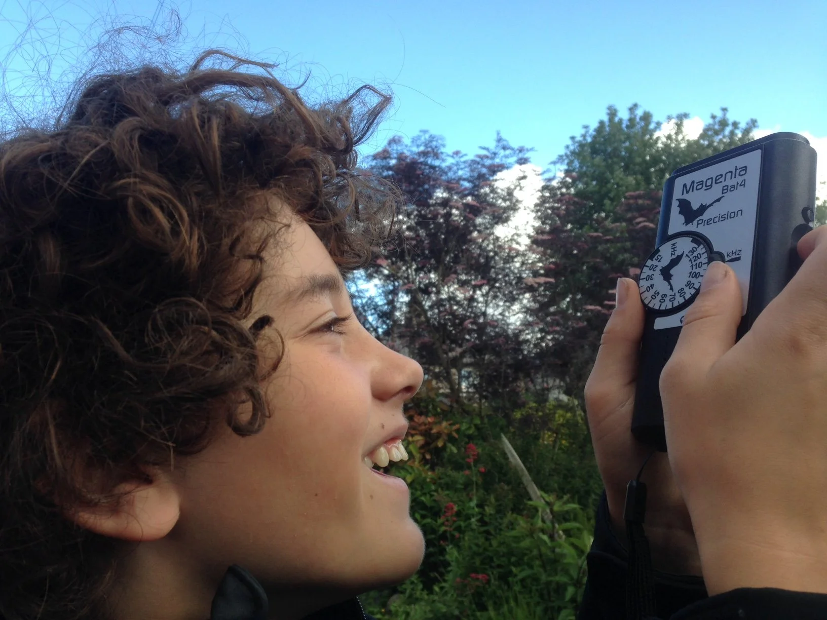 A young boy with curly hair smiling as he looks at a handheld radar device outdoors, with trees and a blue sky in the background.