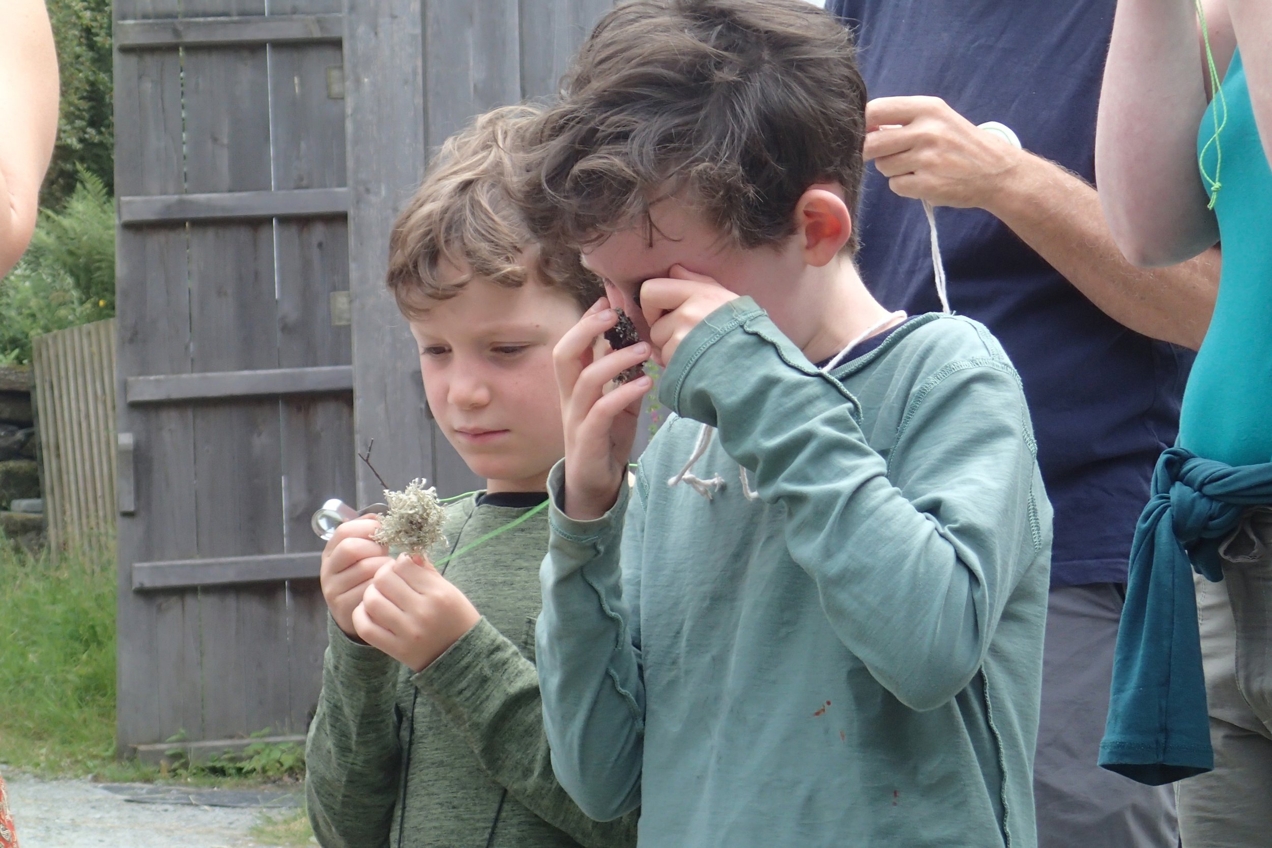 Two young boys examining objects they found outdoors, with a woman in the background. One boy is holding a small plant or flower, and the other boy is holding a small dark object close to his eye, appearing to look through it.