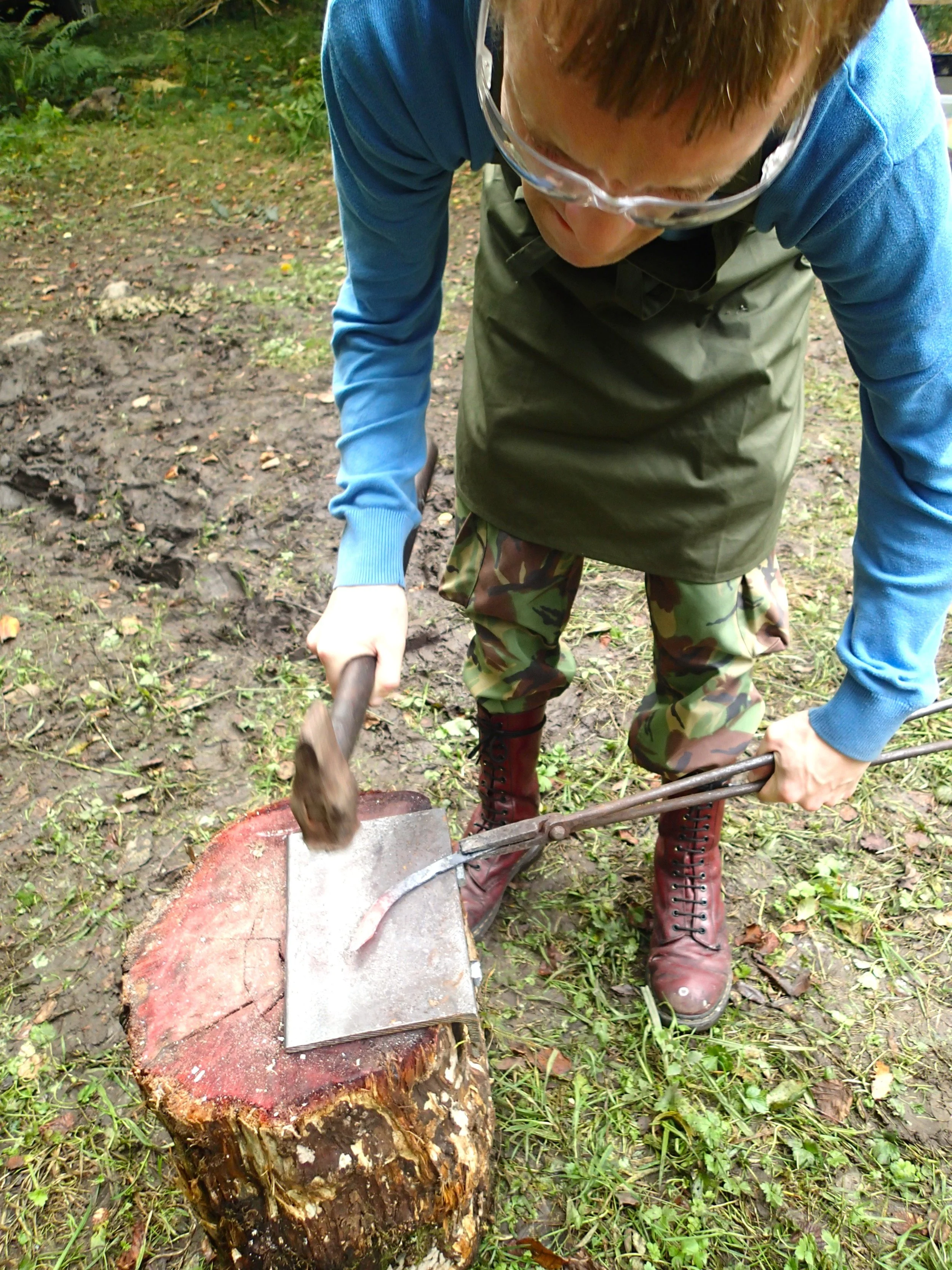 A person wearing camouflage pants, a green apron, and red boots is chopping a large log with an axe outdoors on a muddy ground.