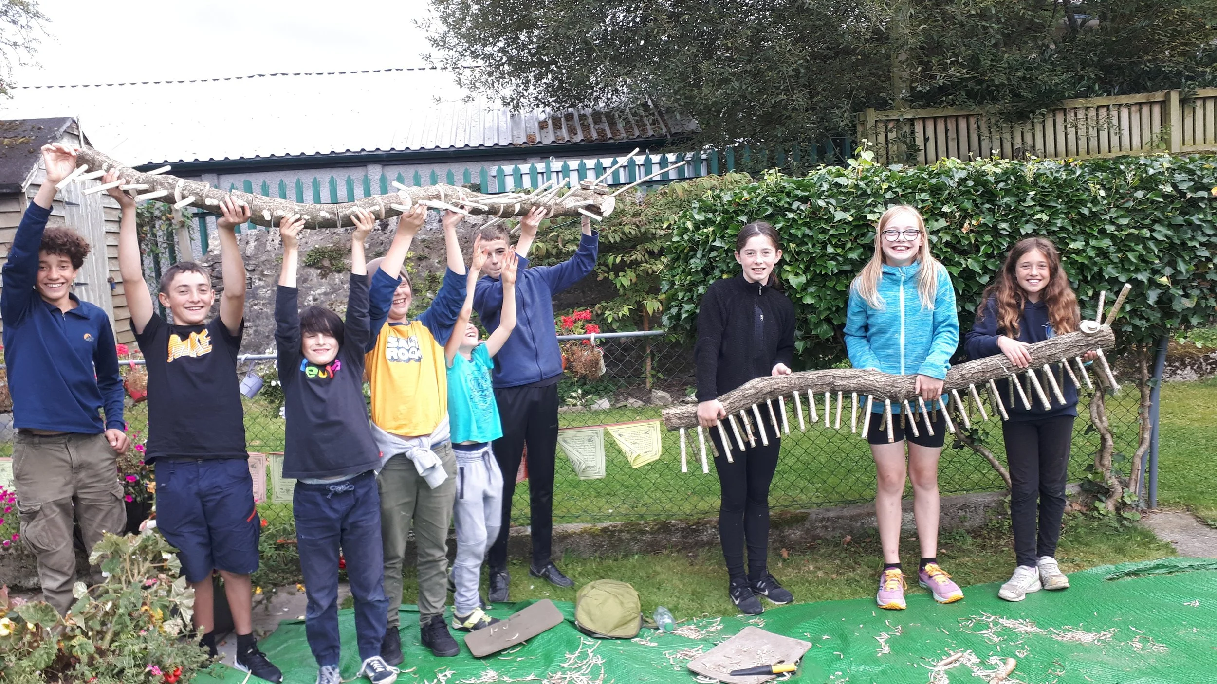 Group of children outdoors holding large wooden sticks assembled as a bridge and a caterpillar, smiling and standing on a green mat in a garden.