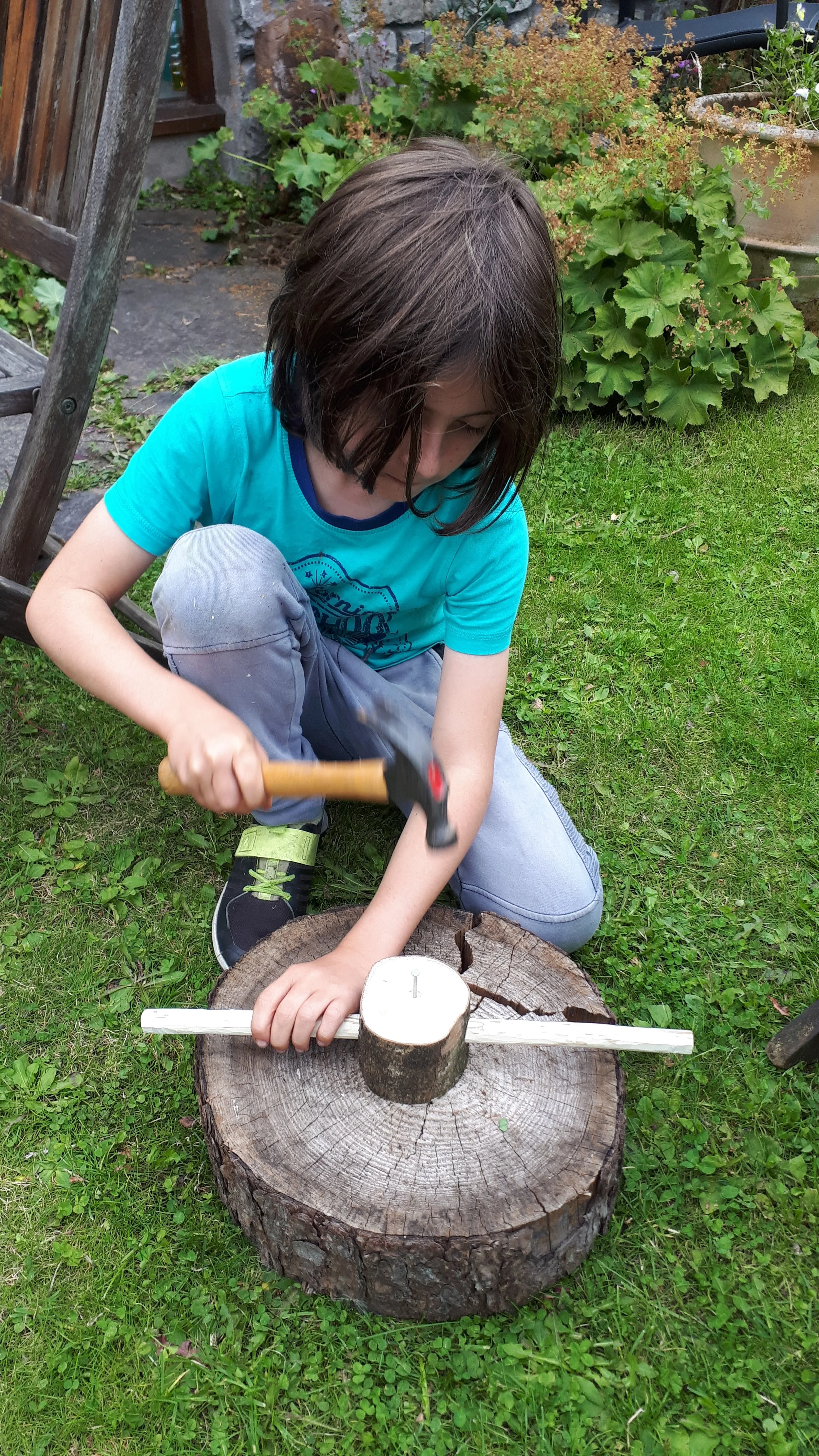 A young boy is outside, sitting on the grass, using a hammer to drive a nail into a round piece of wood placed on a large tree stump. The scene is surrounded by green grass and plants, with a wooden chair and a stone wall in the background.