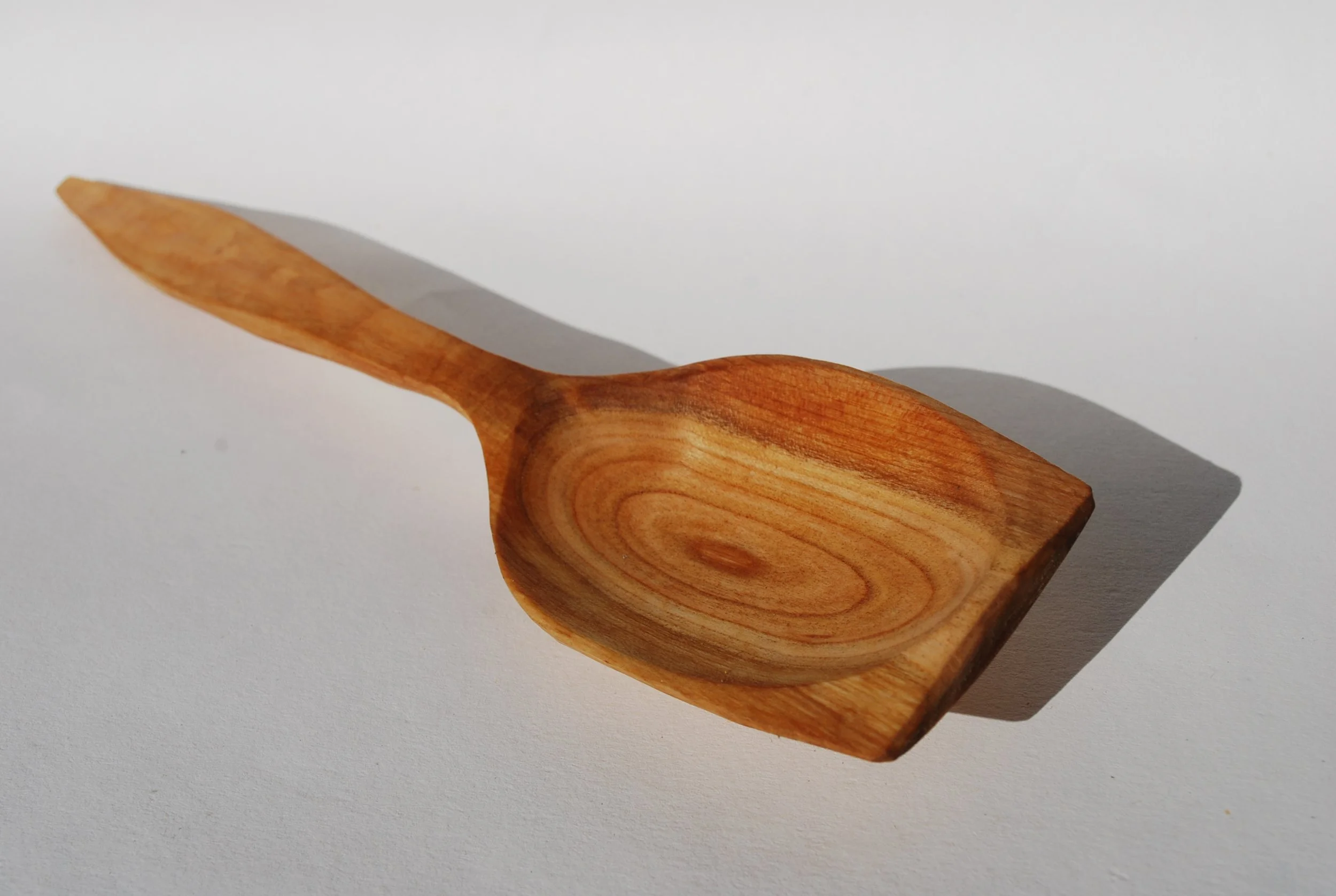 Wooden spoon with a rounded bowl and a tapered handle on a white surface, casting a shadow.